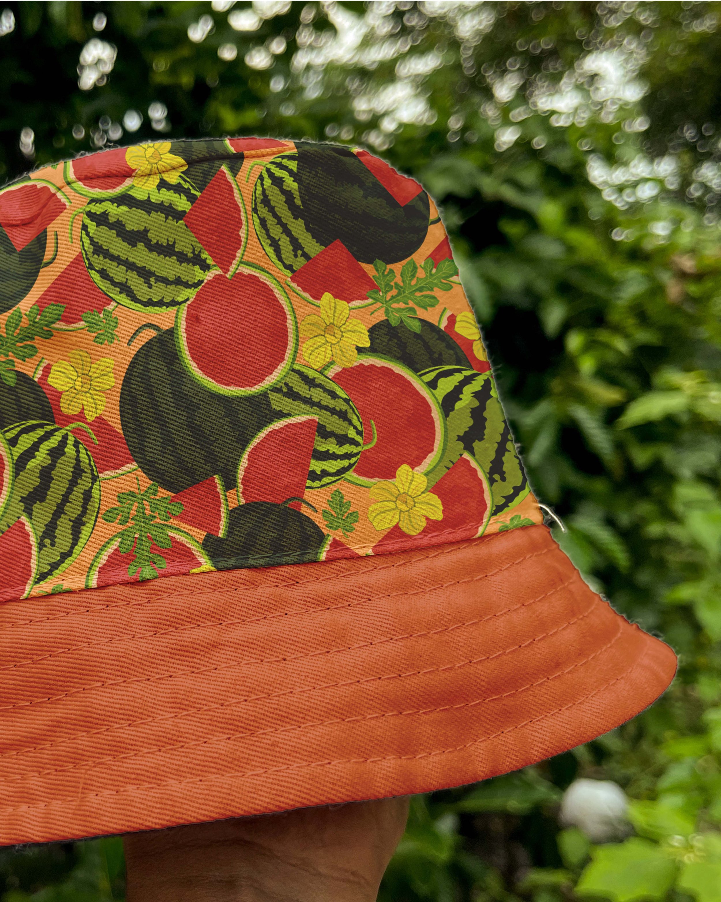 Sombrero naranja con detalles coloridos y un diseño de sandías, hojas y flores, sostenido por una mano en un ambiente exterior con vegetación.