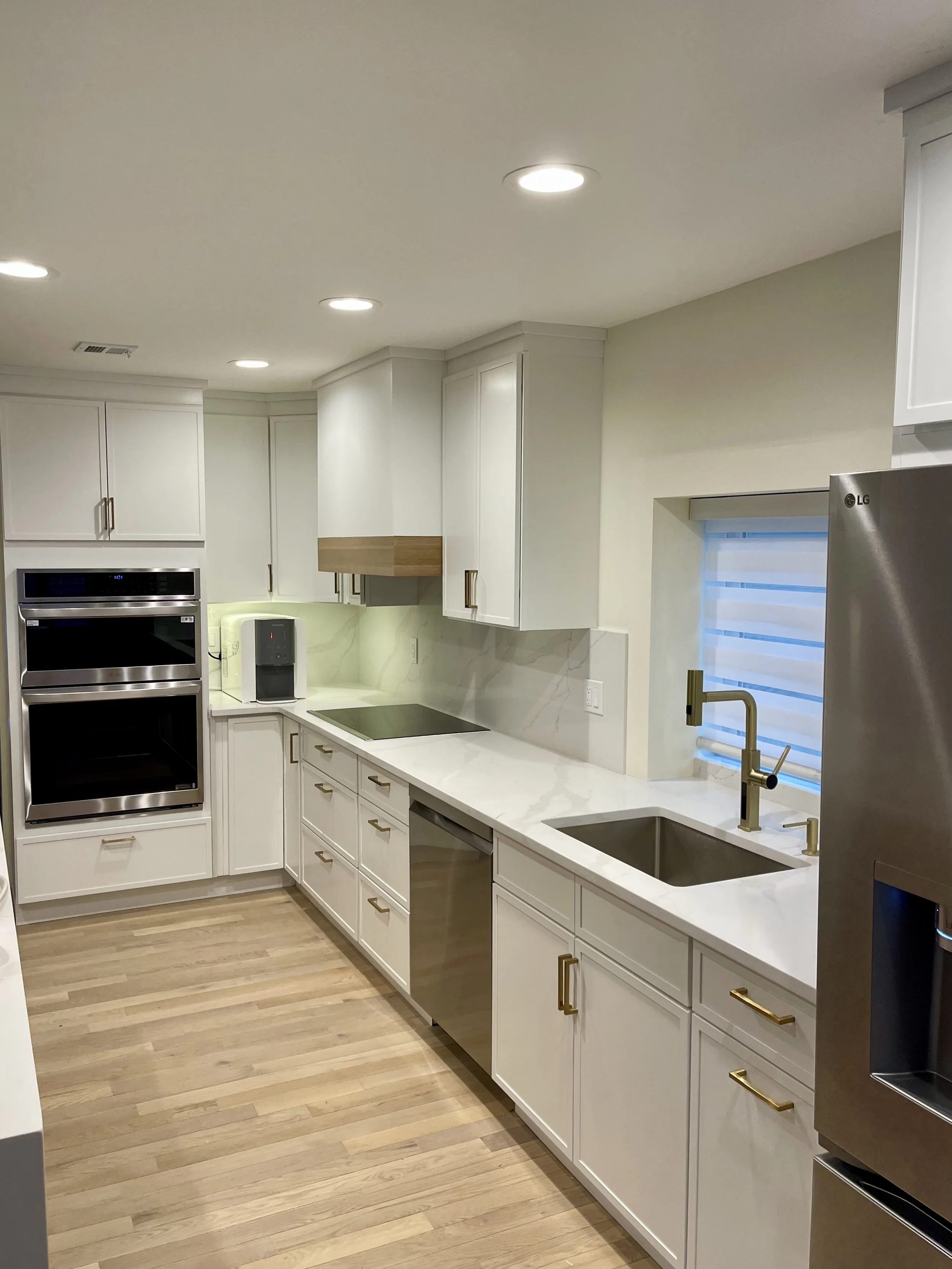 Modern kitchen with white cabinets, gold hardware, stainless steel appliances, a marble countertop, and a window with striped blinds.