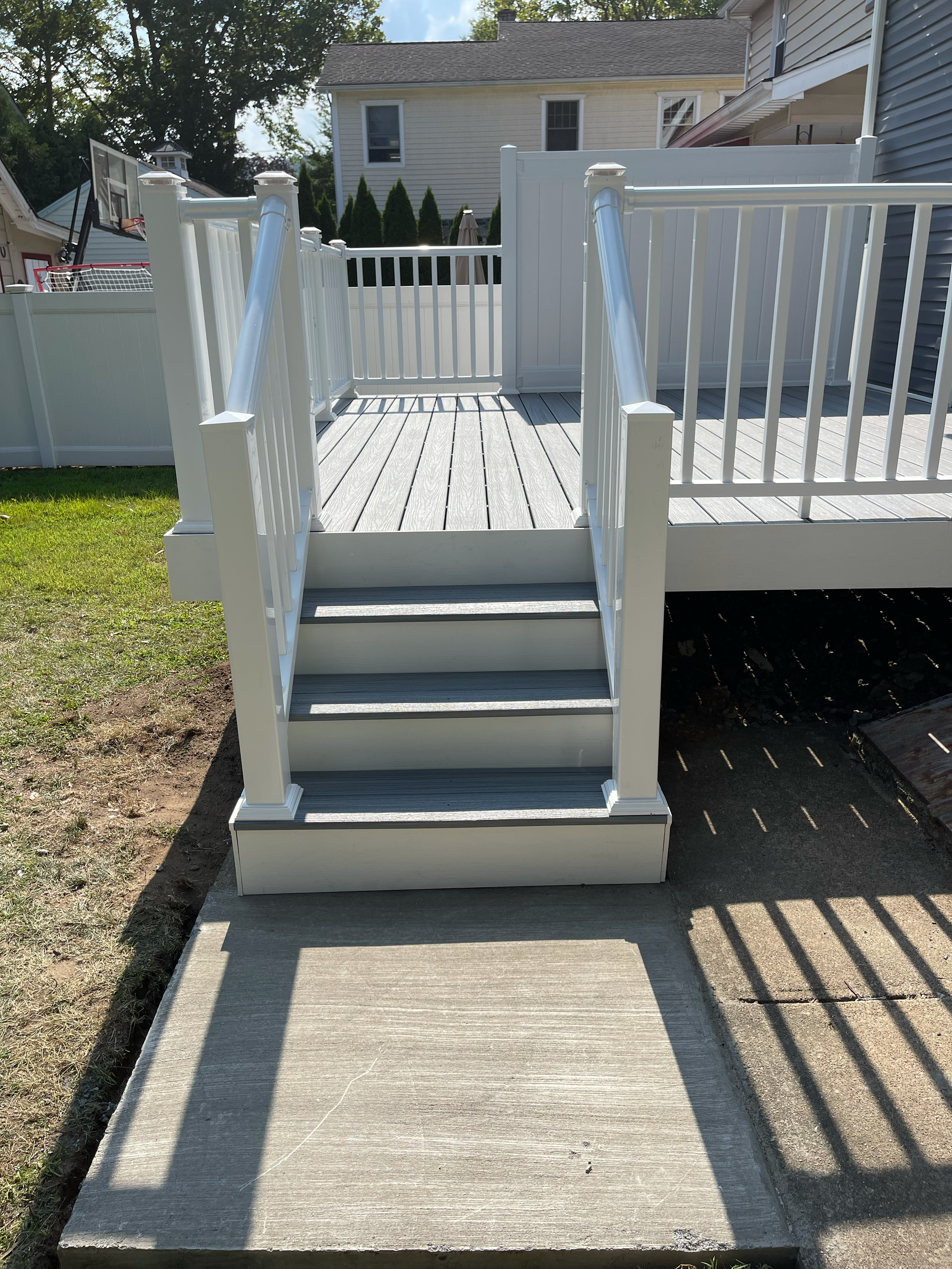 A set of stairs leading up to a backyard deck with white railings, with a concrete walkway in front and a grass lawn to the side.