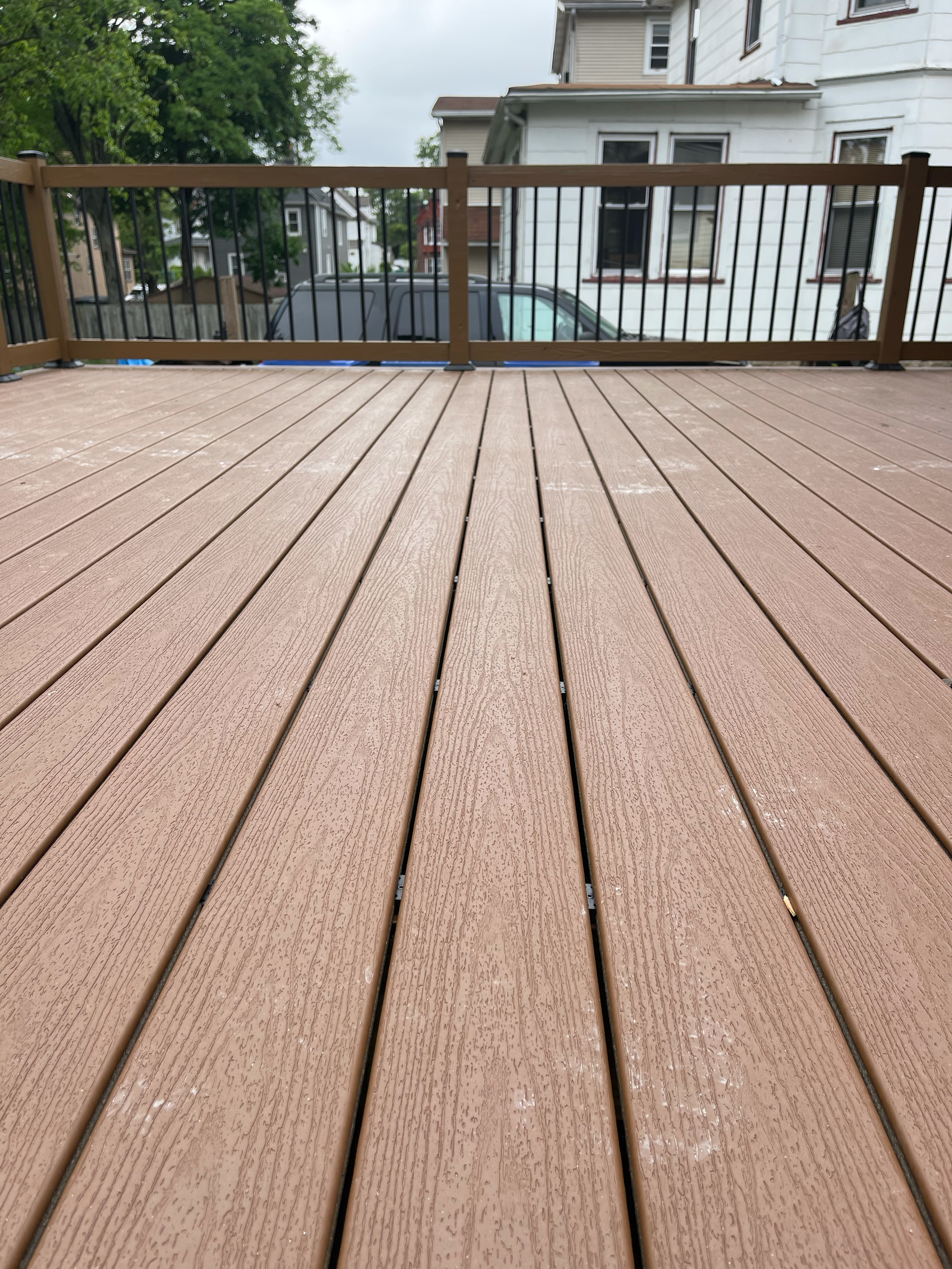 View of a newly built wooden deck with a brown railing, outdoor residential area in the background, with houses and a gray vehicle parked nearby, overcast sky.
