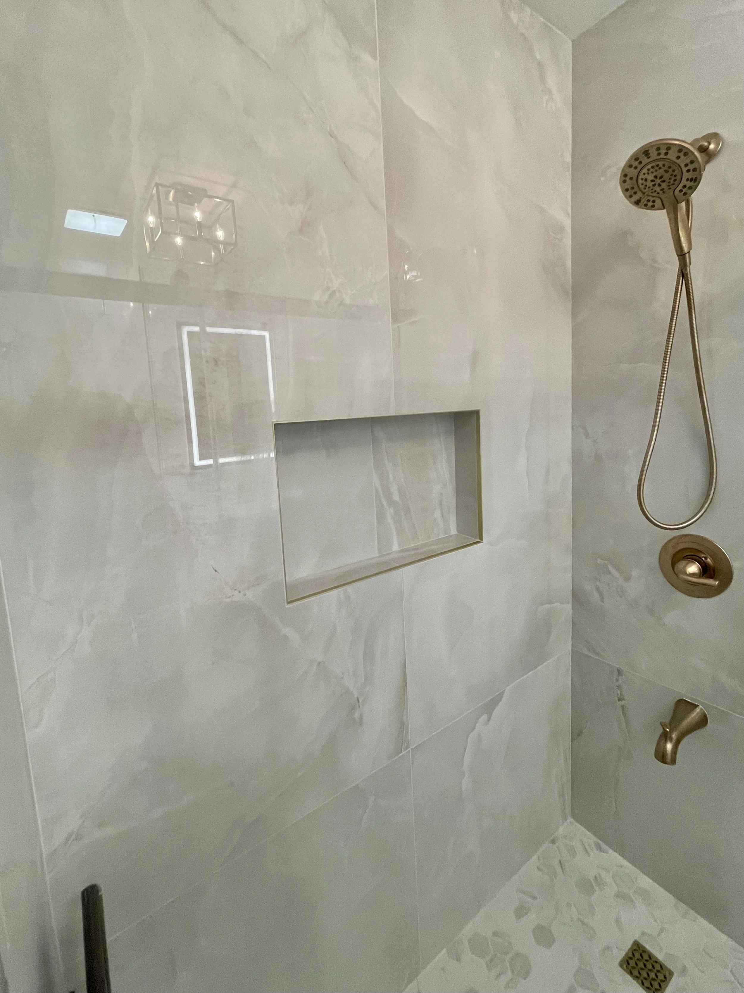 A modern shower with large beige marble tiles, a built-in niche for toiletries, a brass shower head with a flexible hose, and a brass water valve. The floor has small hexagonal tiles and a drain.
