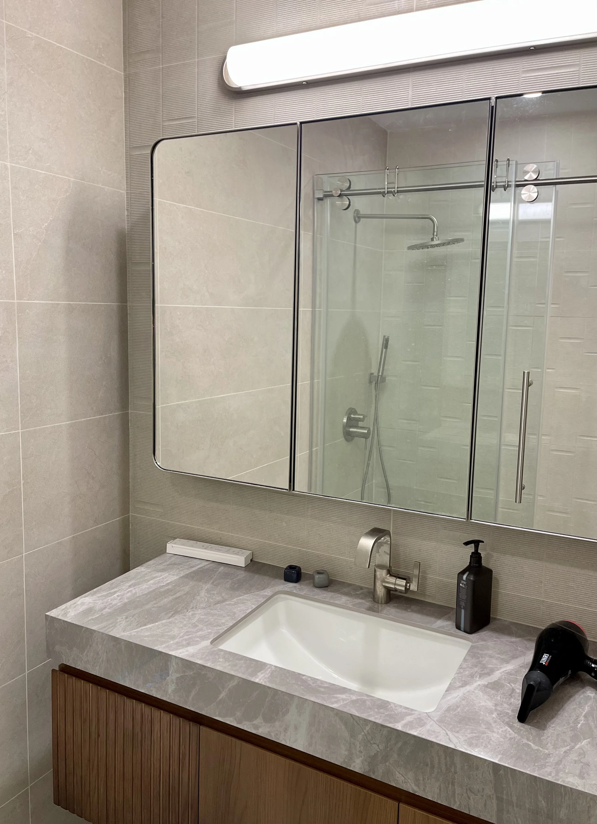 Bathroom vanity with a rectangular sink, black soap dispenser, and a hairdryer on a marble countertop. A large mirror with a light strip above and a glass shower enclosure are visible in the background.