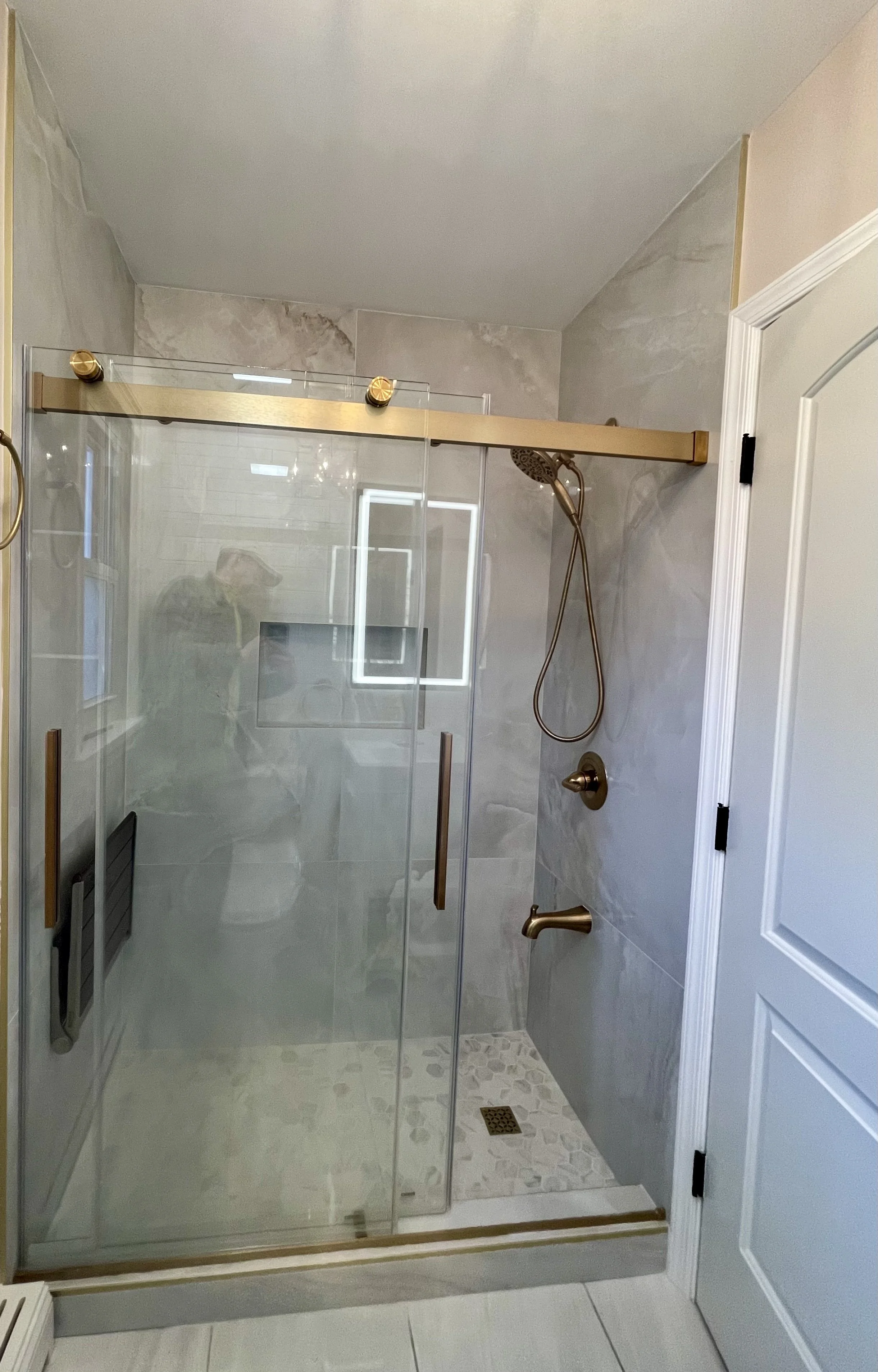 A modern walk-in shower with clear glass sliding doors and gold fixtures, including an overhead showerhead, a handheld shower, and a gold handle on the door. The shower has gray marble walls, a pebble stone pattern on the floor, a shower niche, and a