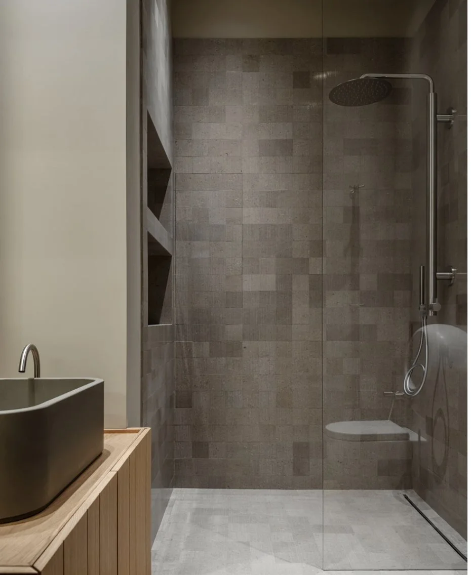 A modern bathroom with a walk-in shower enclosed by a glass partition, tiled walls, a ceiling-mounted showerhead, a wooden vanity with a black vessel sink, and built-in shelving in the shower area.
