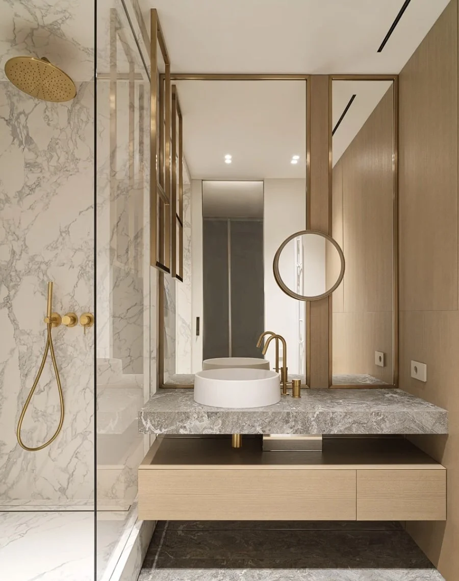 Modern bathroom with a marble shower wall, gold fixtures, a floating wooden vanity with a marble countertop, a vessel sink, a round mirror, and minimalistic lighting.