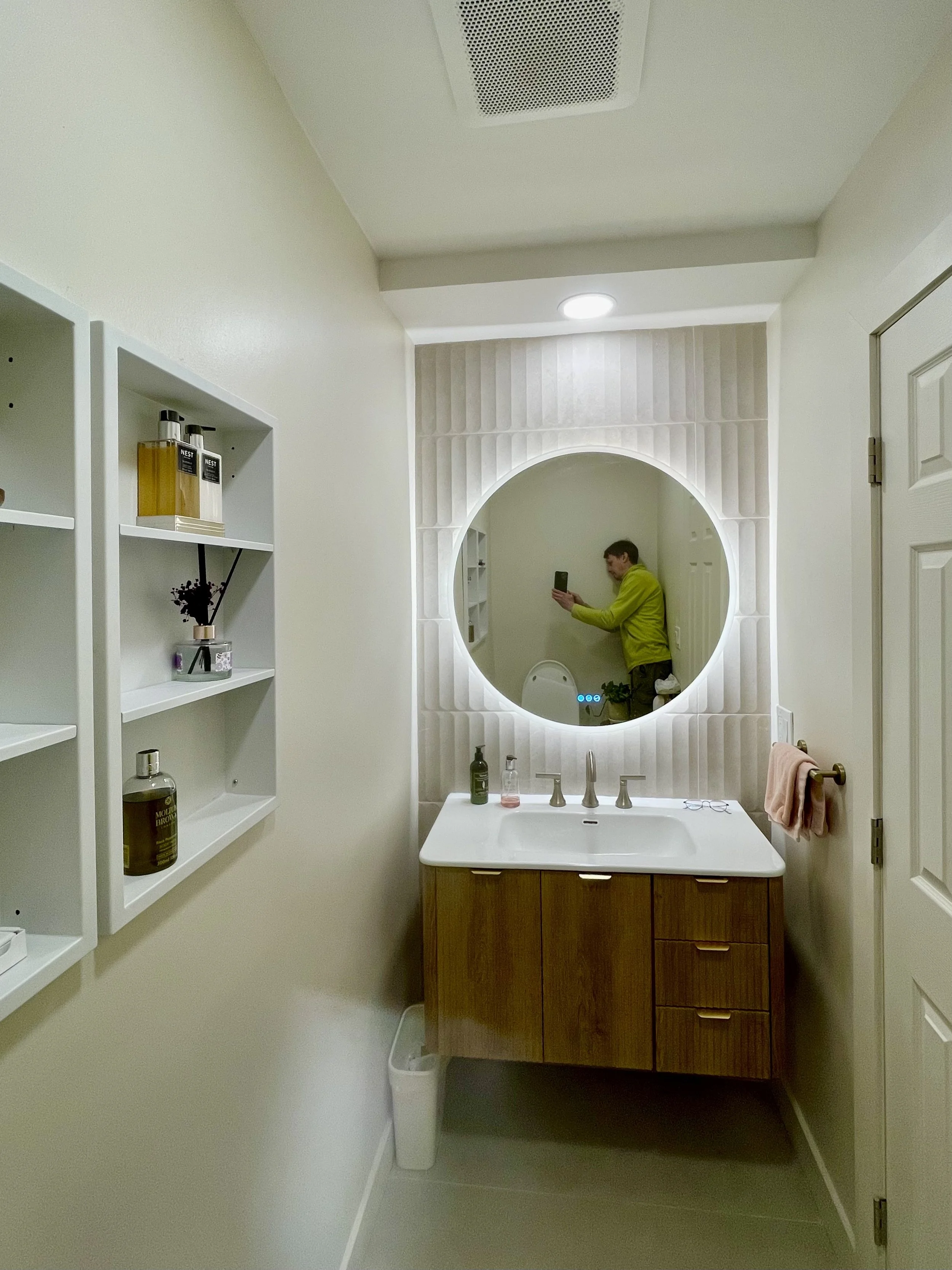 Modern bathroom with a round mirror, wooden vanity, and white walls; a person taking a selfie is reflected in the mirror.