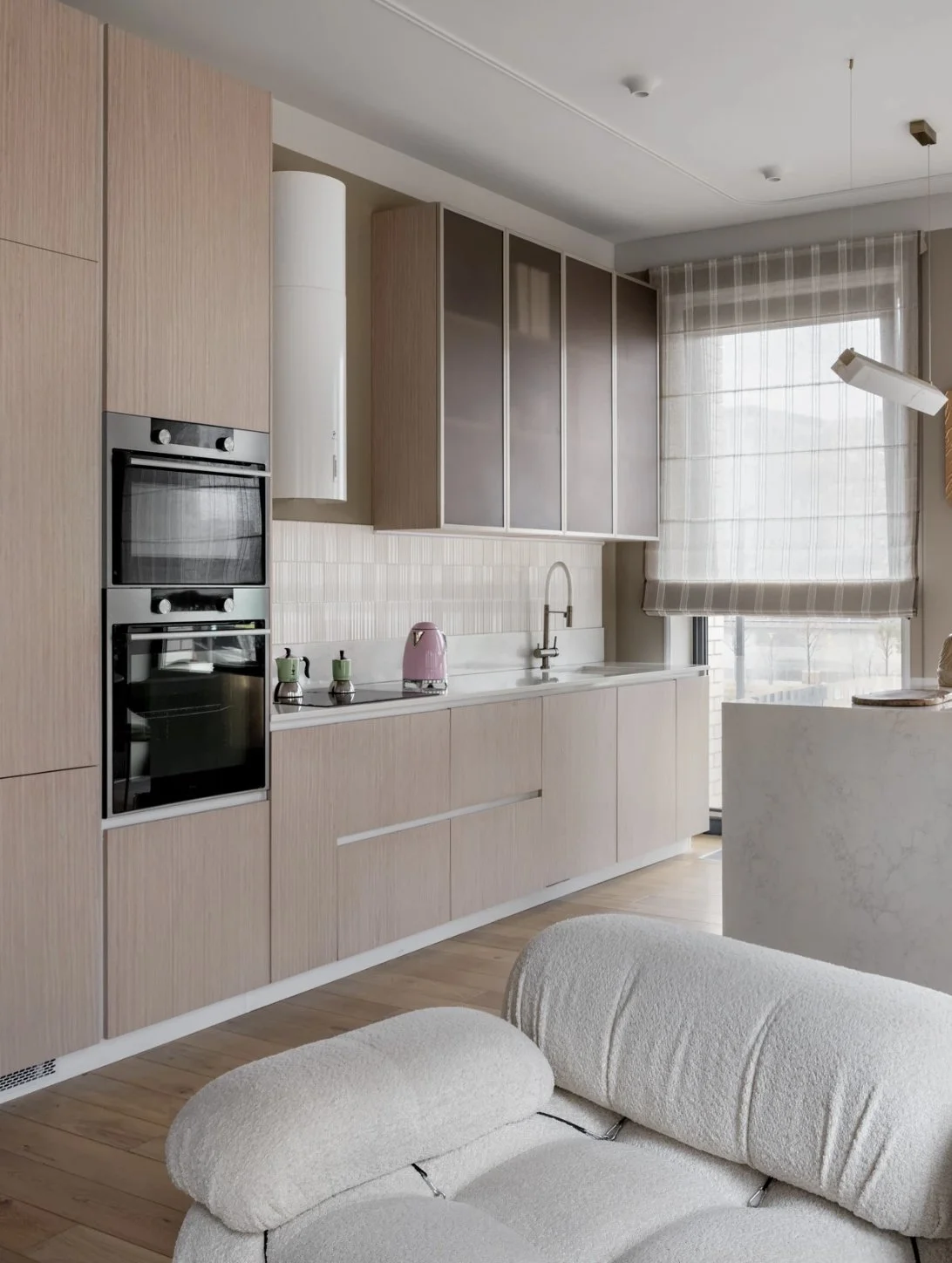 Modern kitchen with light wood cabinets, white countertops, a pink kettle, and a large window with striped blinds.