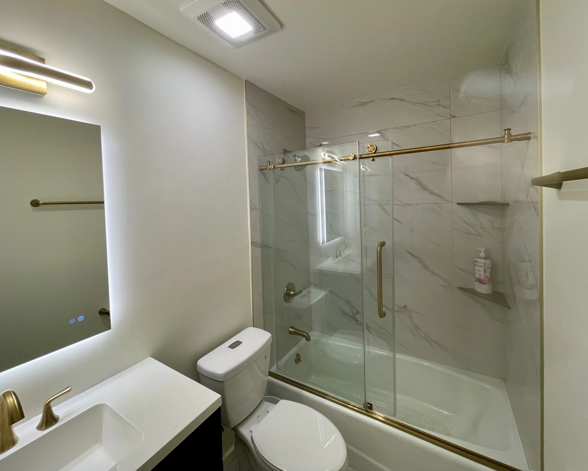 Modern bathroom with a glass-enclosed shower with gold fixtures, a white toilet, a sink with a gold faucet, and a mirror with built-in lighting.