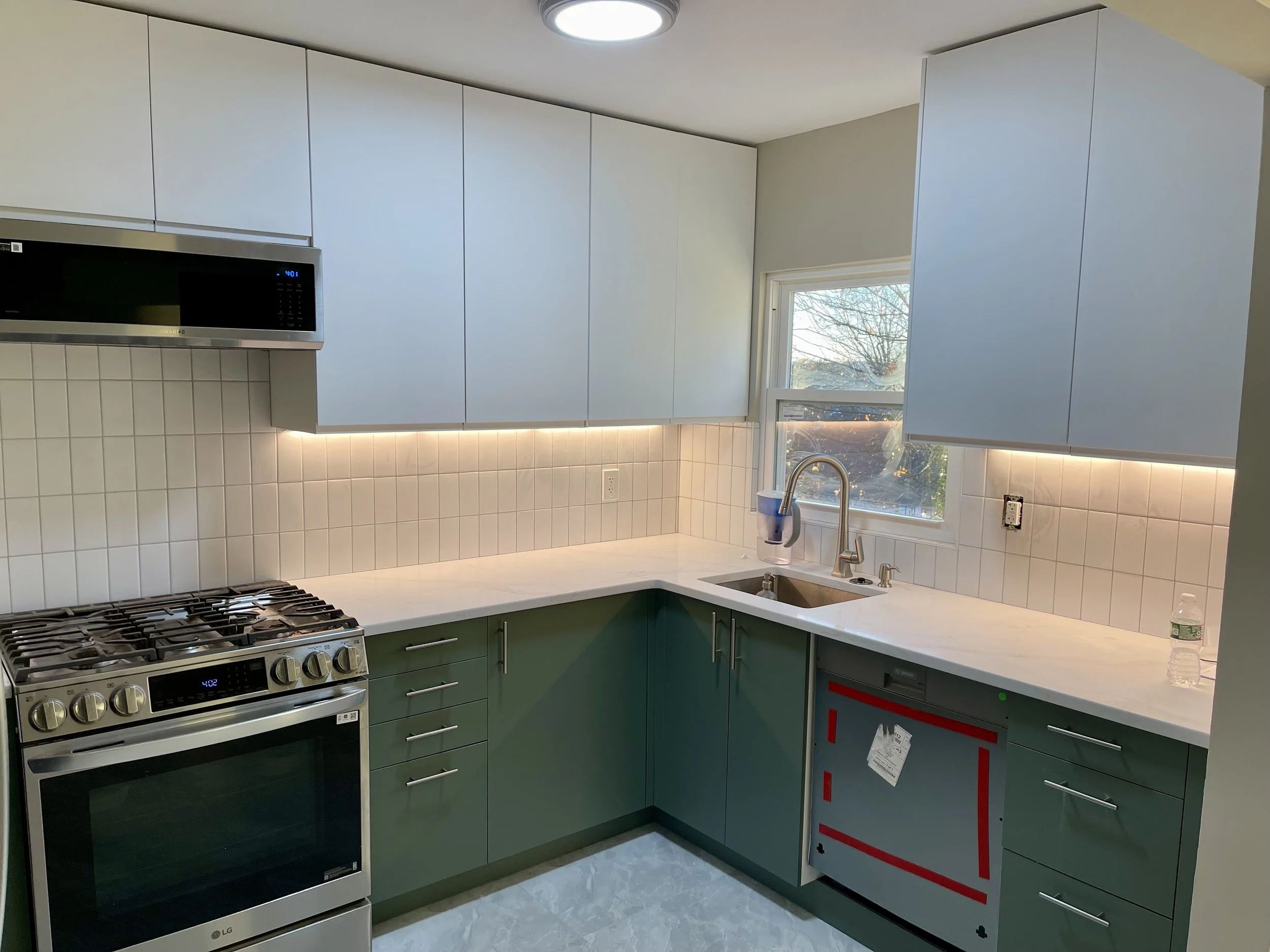 Modern kitchen with green lower cabinets, white upper cabinets, a white countertop, beige tiled backsplash, stainless steel stove, microwave, and a window above the sink showing outside trees.
