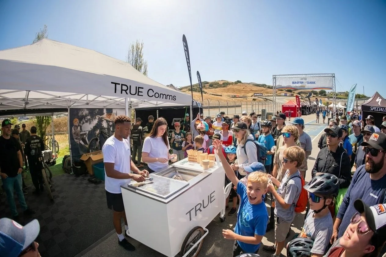 TRUE Comms branded ice cream cart surrounded by a crowd of event goers at the Sea Otter Classic cycling event, with a TRUE Comms branded tent visible in the background.