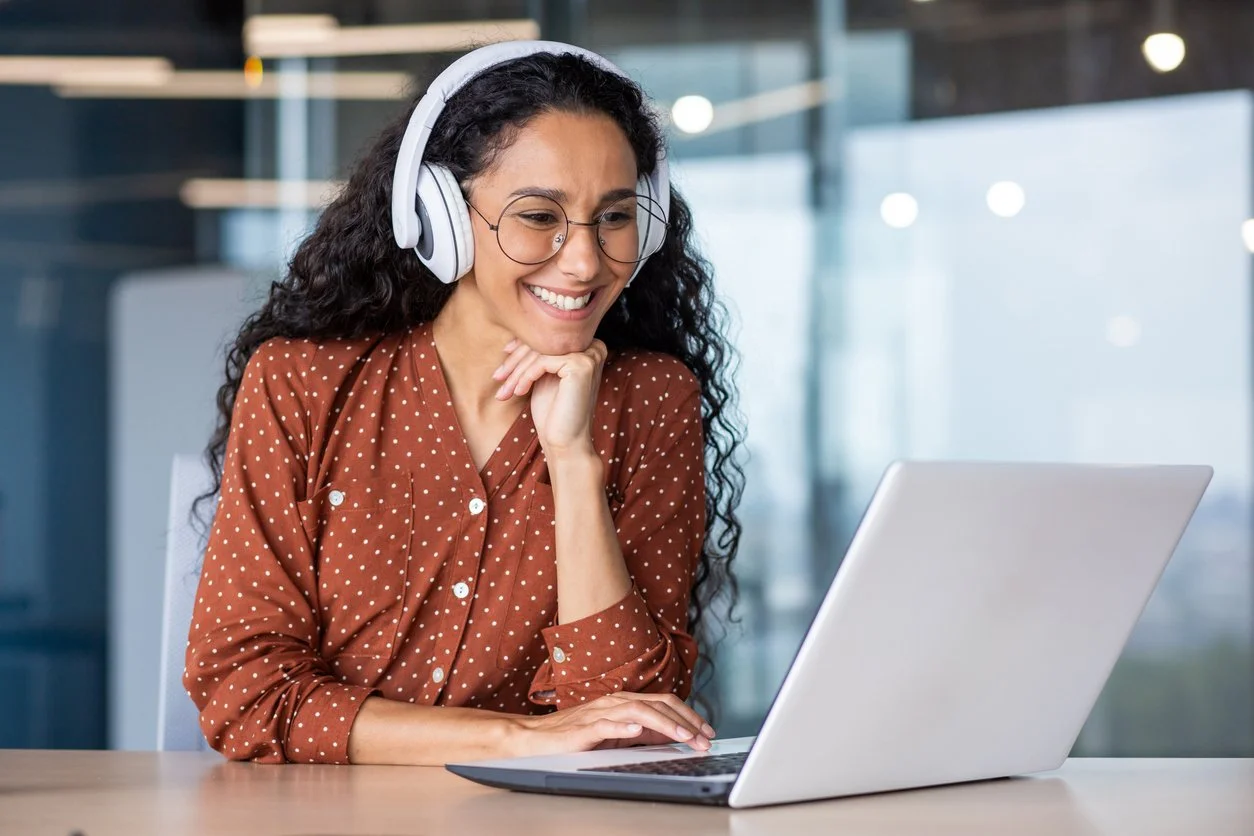 A woman with curly dark hair wearing glasses and a brown shirt with white polka dots, smiling while looking at her laptop and wearing white headphones in a modern office environment.
