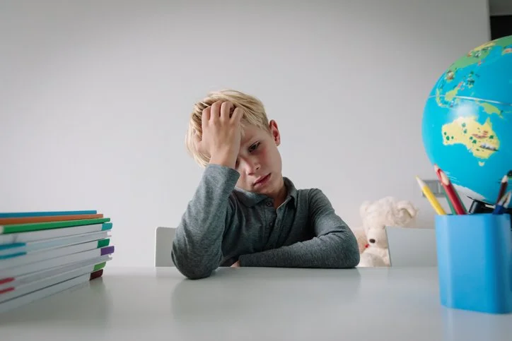 A young boy sitting at a desk with a stack of books, a globe, and a teddy bear, looking overwhelmed and holding his head.