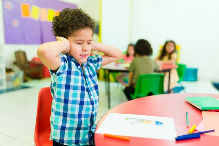 A young boy in a blue plaid shirt holding his ears and wincing in discomfort in a classroom with other children in the background.