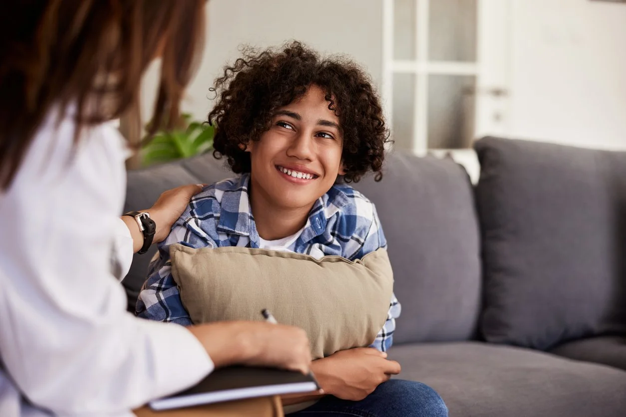 A young boy with curly hair smiling, sitting on a couch, talking to a woman, who is taking notes.
