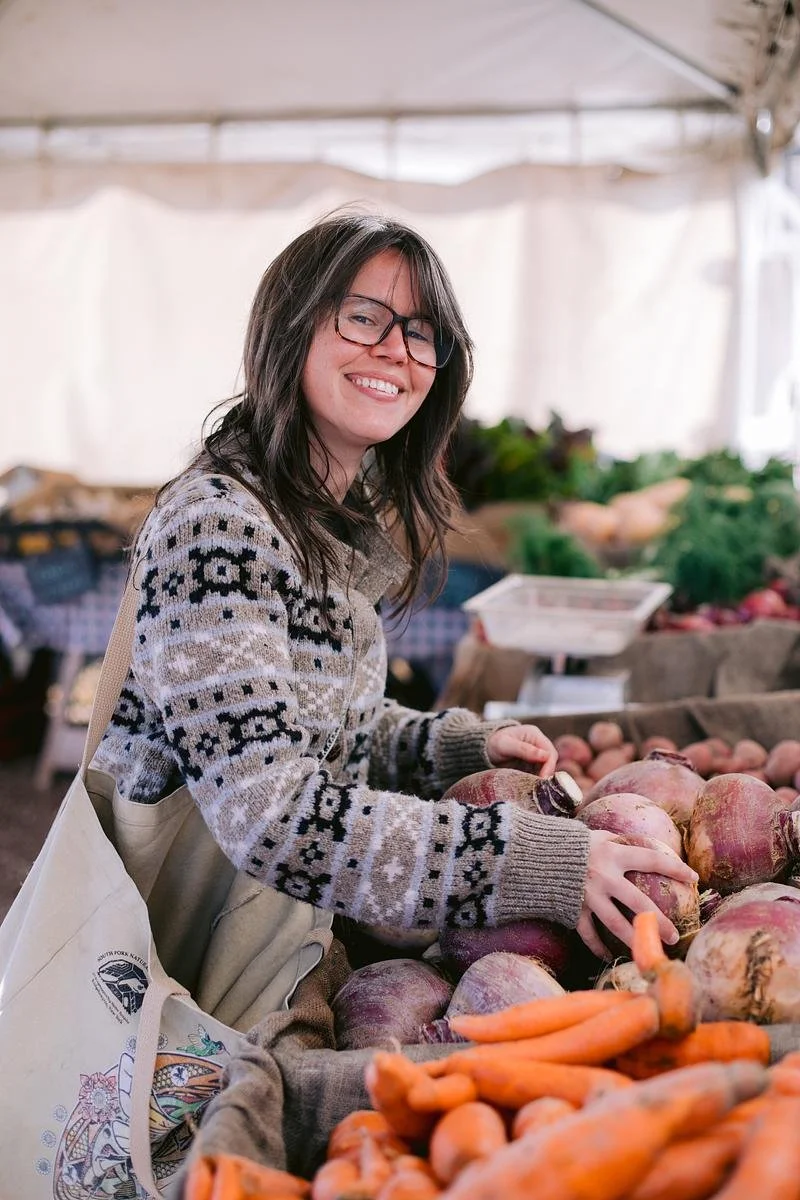 A table a red and white checkered covering runs at an angle from the foreground to the background. It's covered in bins lined with burlap sacks and filled with a variety of produces. Two eople can be seen reaching their hands into the bins.