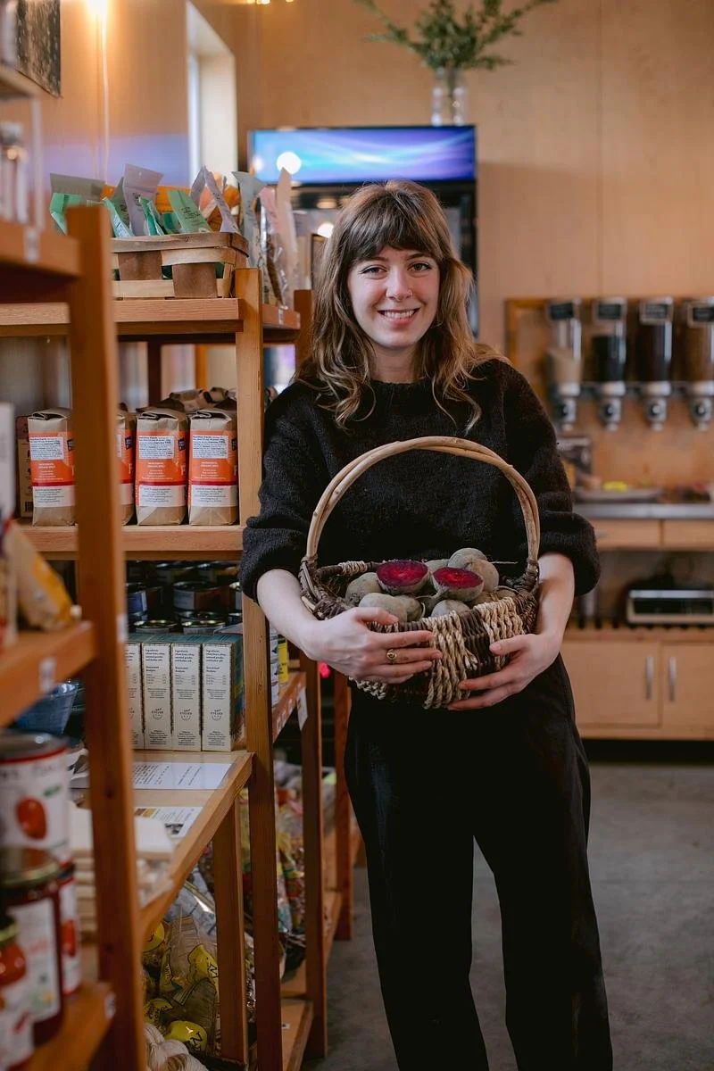 A Farm Store employee wearing black pants and a black sweater leans against a shelf full of pantry items while holding a woven basket full of red beets.