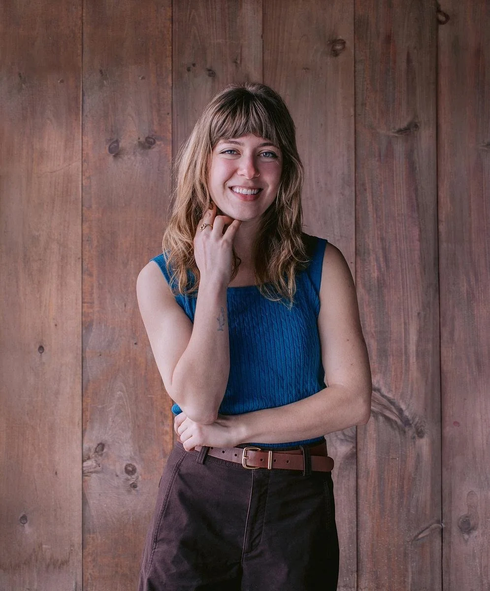 Anna, wearing a sleeveless, blue top and brown pants, crosses her arms with her hand under her chin and stands in front of a wood-paneled wall.