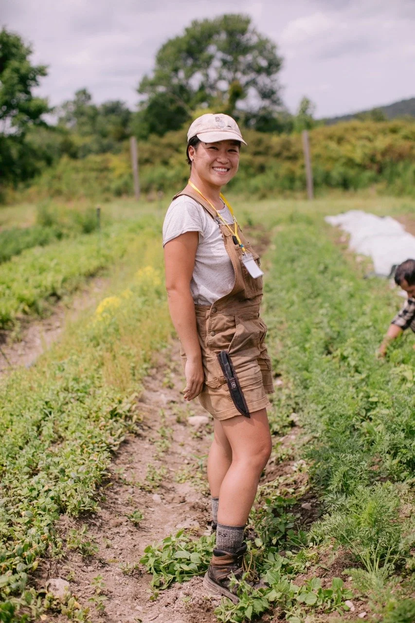 A Farmer wearing brown short overalls stands sideways with their head turned to the camera smiling. They are standing between two rows of plants in a field. Another farm can just be seen crouching in the background reaching out towards the plants.