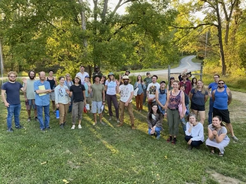 A group of people stand oustide on a grassy area with a few trees and windy paved road behind them. The sun is showing through the trees and casting long shadows.