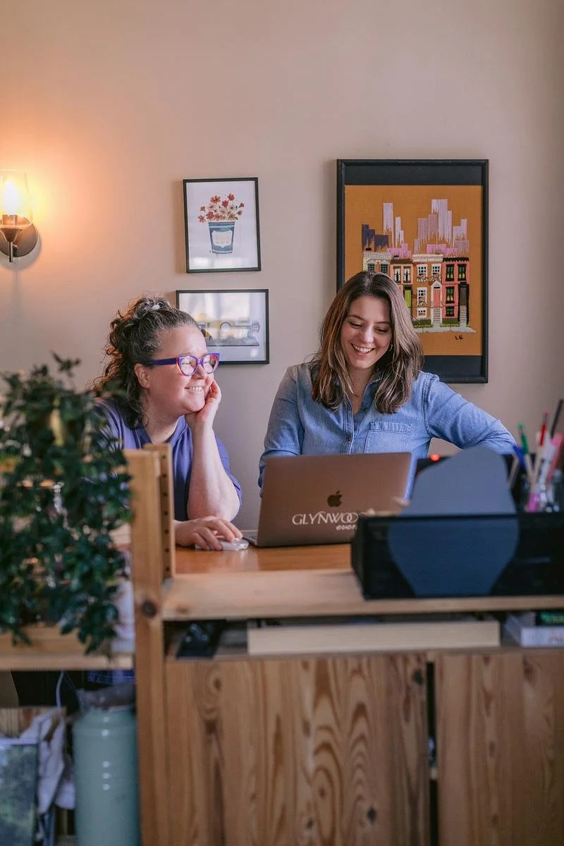 Two people stand behind a desk looking at a laptop screen.