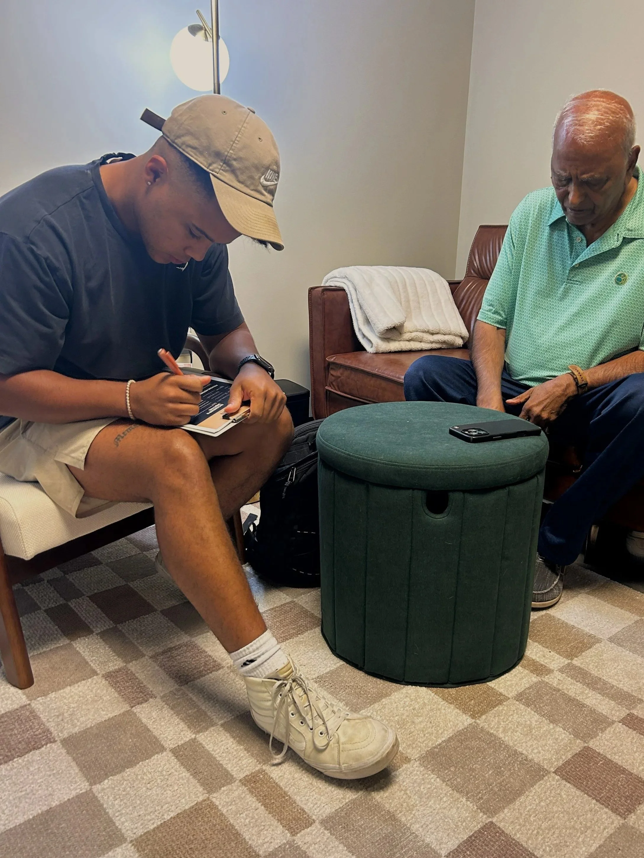 A young man in a beige cap writing in a notebook while sitting on a chair next to an older man with gray hair wearing a light green polo shirt, sitting on a brown leather chair with a small green ottoman between them, in a waiting room with beige carpet and a beige wall.