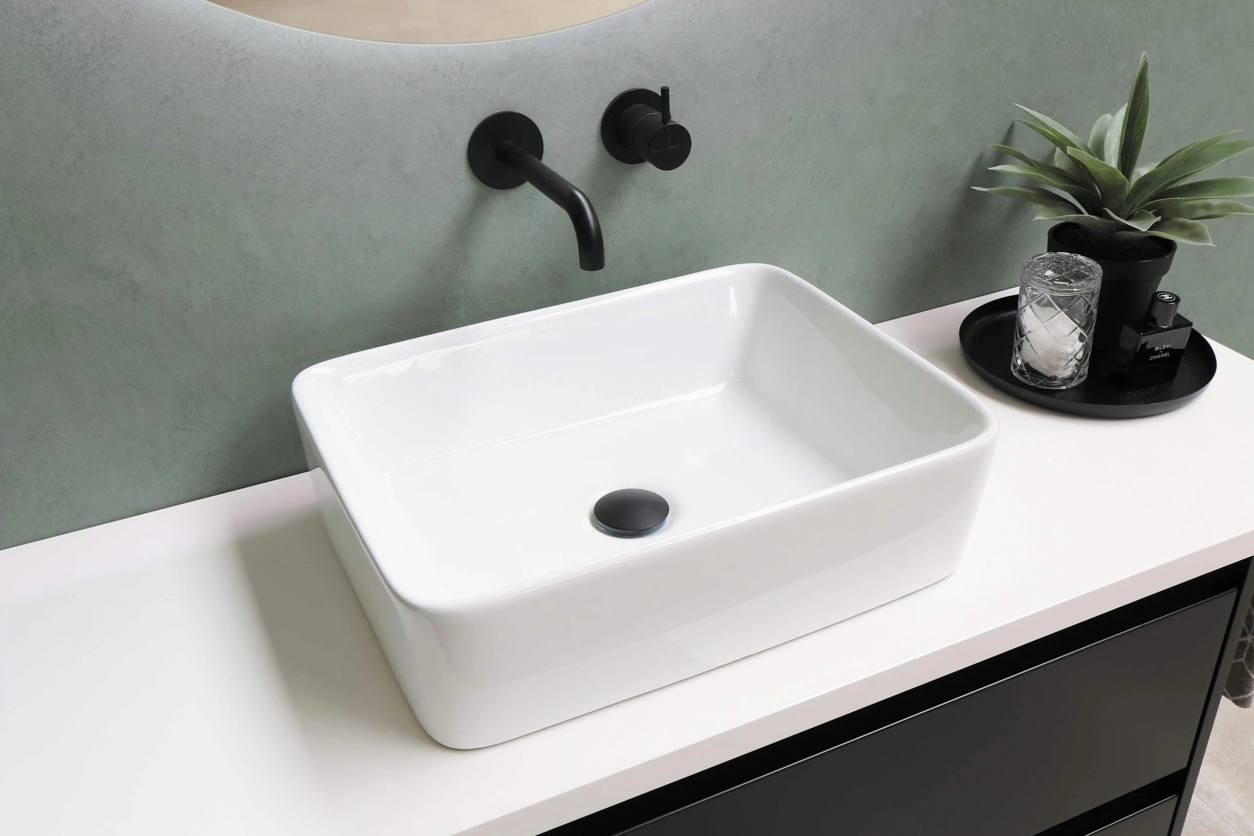 Modern bathroom sink with a black matte faucet, a small black tray holding a glass, a black perfume bottle, and a potted plant, set on a white countertop with a green textured wall background.