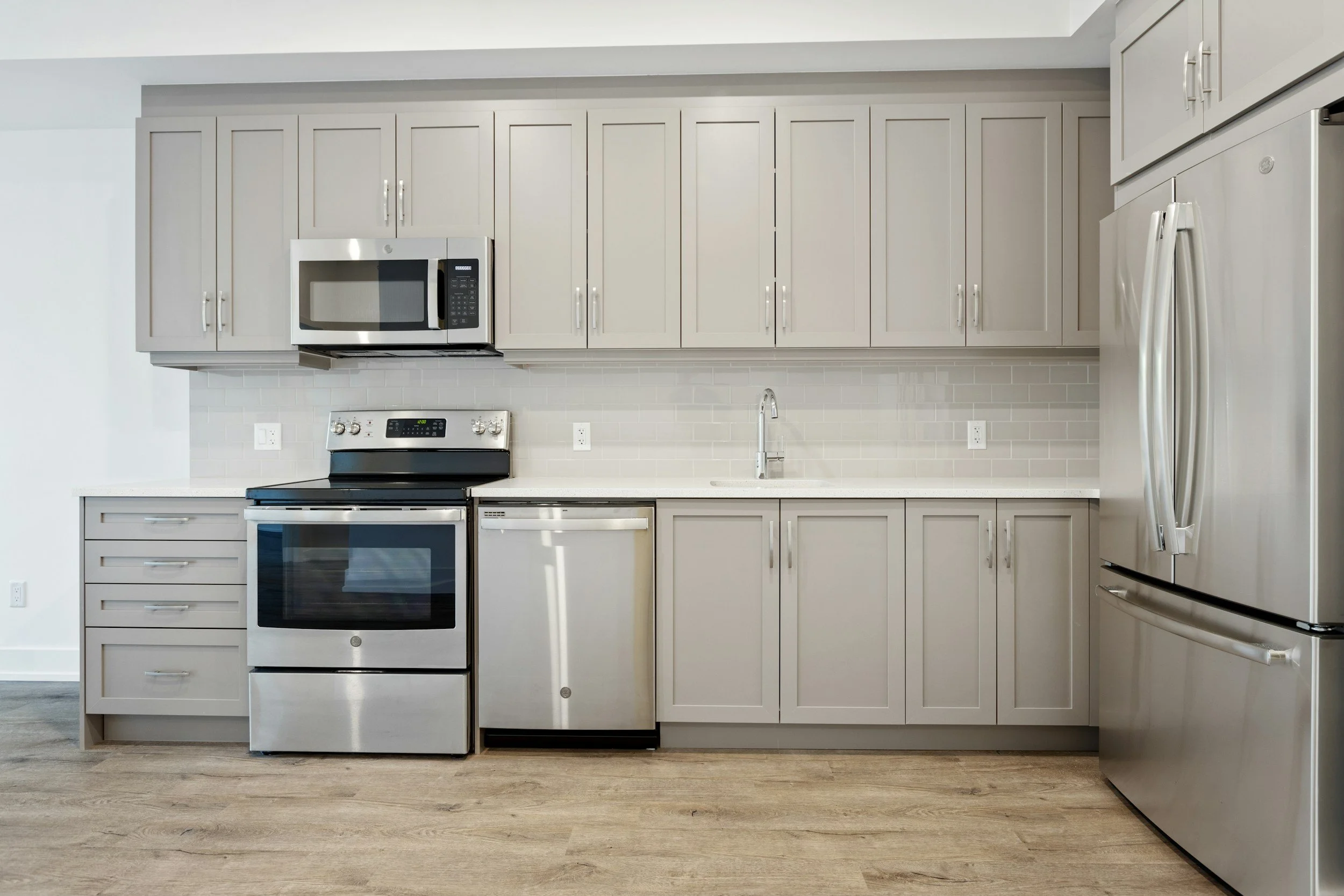 Modern kitchen with stainless steel appliances, light grey cabinets, white backsplash, and light wood flooring.