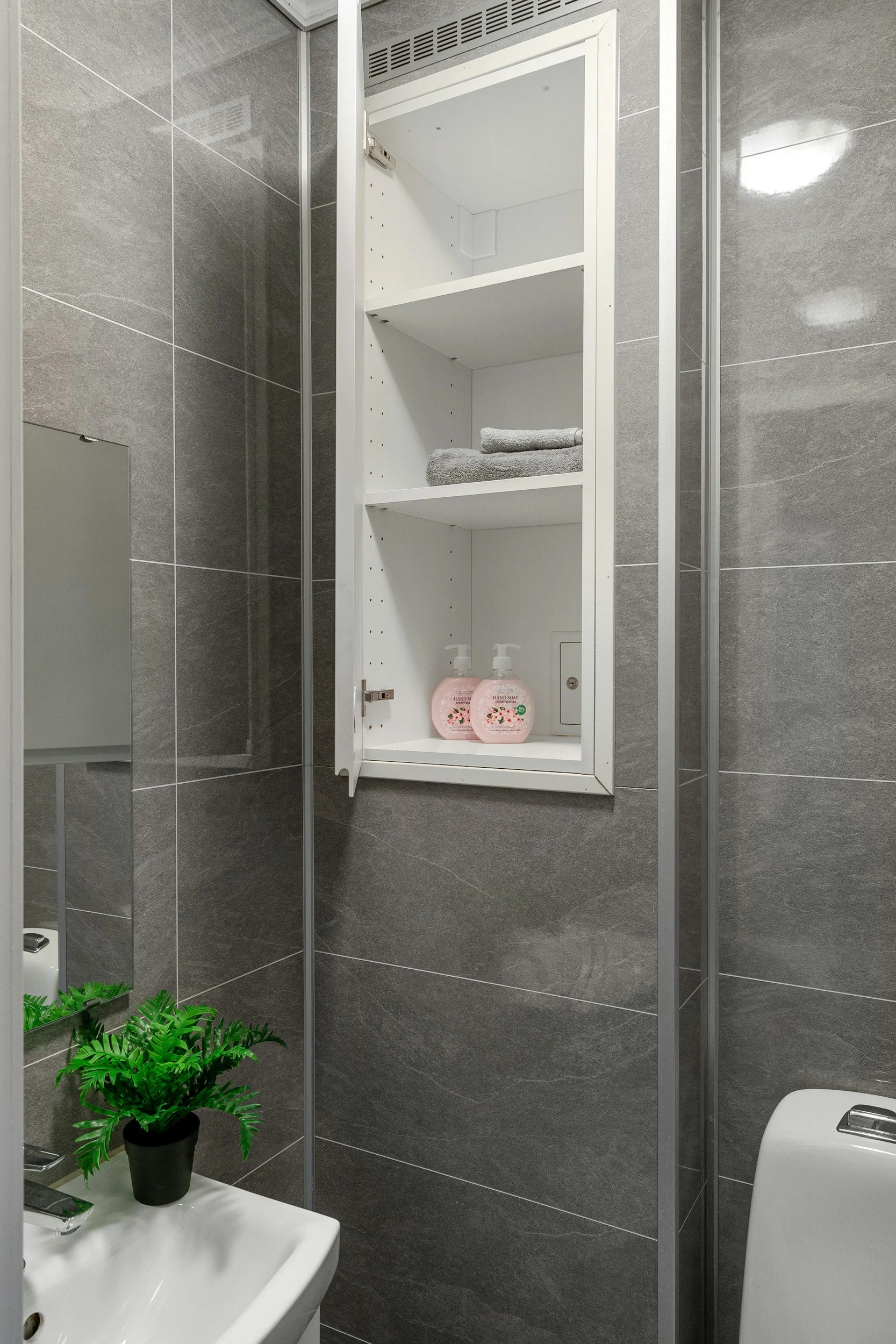 Bathroom with gray tiled walls, white medicine cabinet with towels and pink hand soap bottles inside, a small white sink with a green plant, and a toilet.