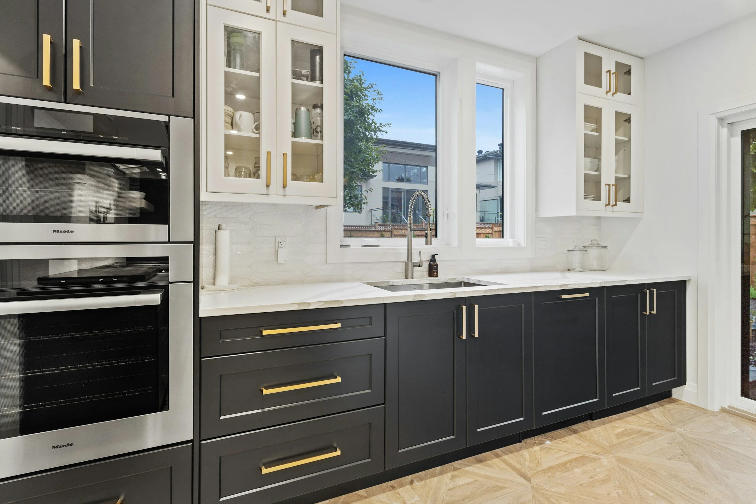 Modern kitchen with black lower cabinets, white upper cabinets with glass doors, stainless steel appliances, white marble countertop, and a window above the sink