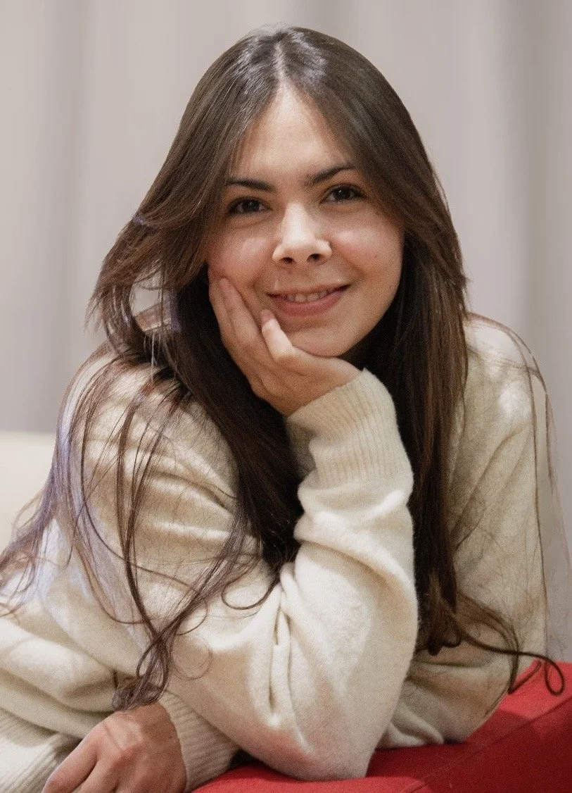 A young woman with long brown hair smiling and resting her face on her hand, sitting indoors against a neutral background.