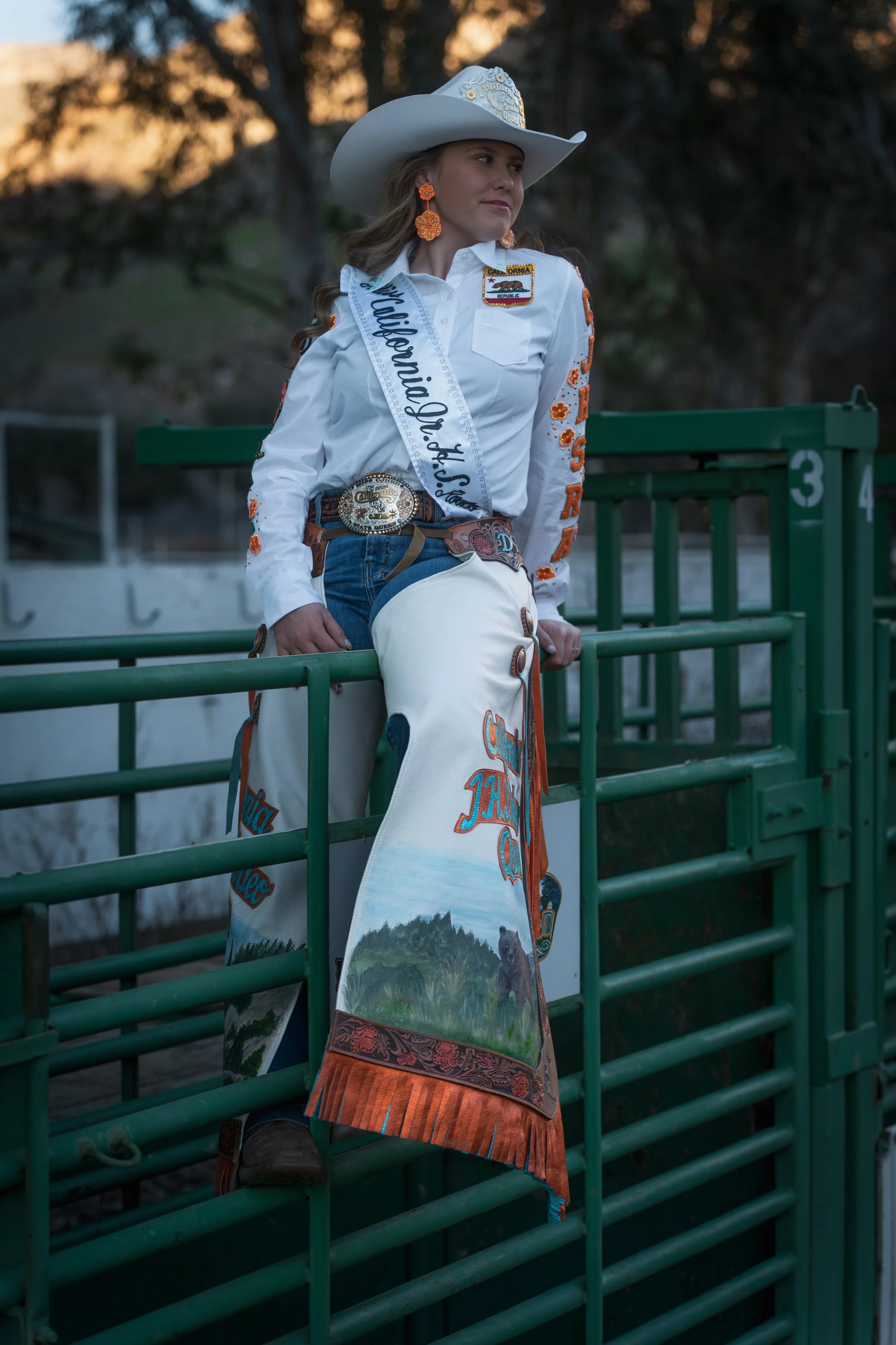 A woman dressed as a cowgirl, wearing a white cowboy hat decorated with badges, orange earrings, a white western shirt with orange embroidery, a belt with a large buckle, and beige chaps with colorful designs, leaning on a green metal fence at a rode