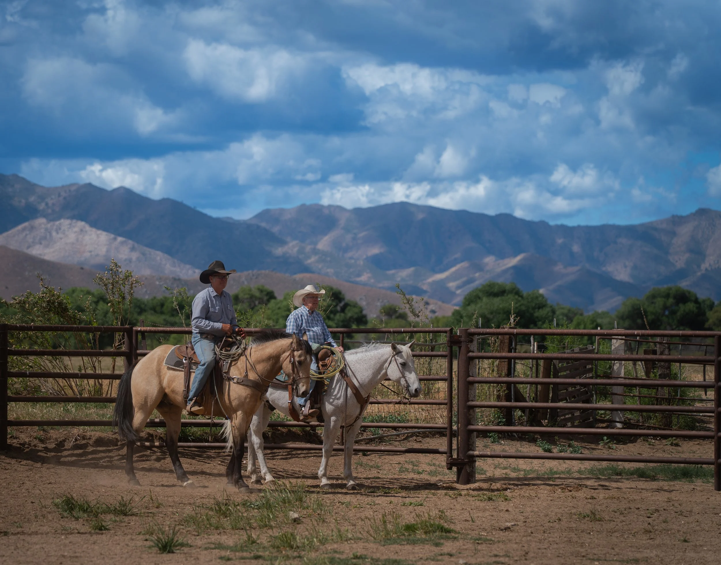 Two men riding horses in an enclosed area with mountains and cloudy sky in the background.