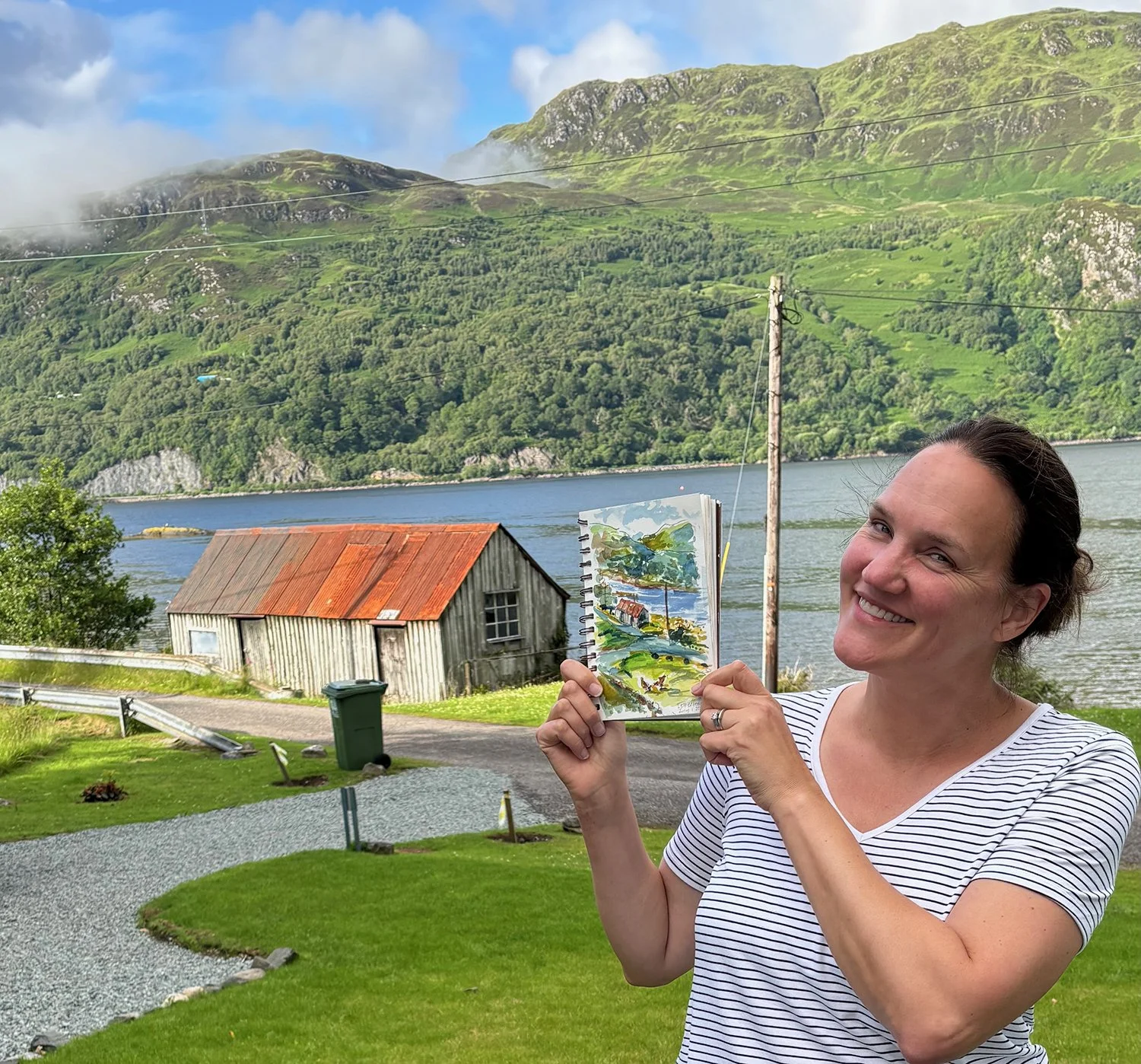 Kathryn holding up a finished painting in front of a beautiful lake in the Scottish Highlands