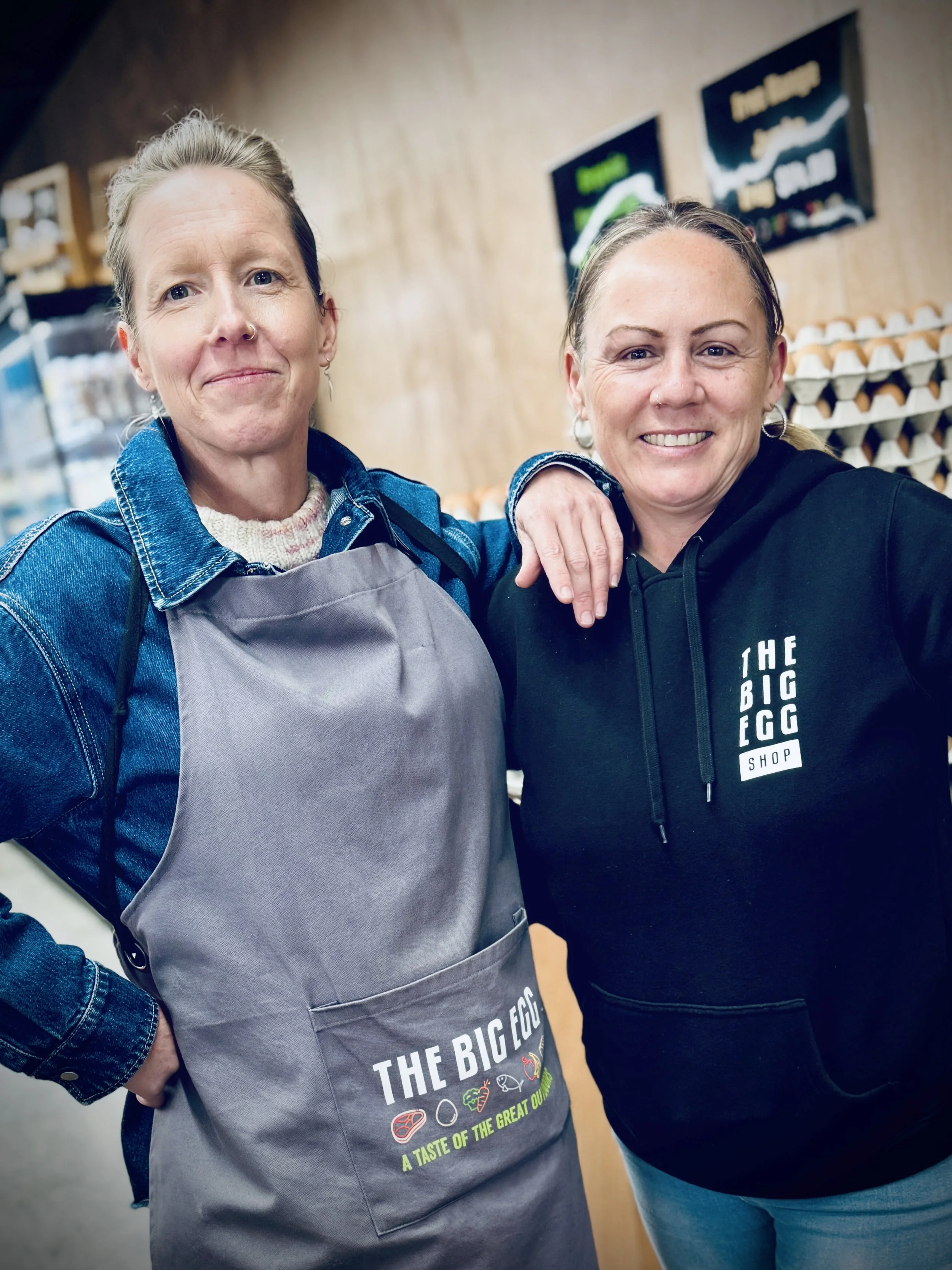 Two Big Egg Shop employees, with one wearing a grey grocery apron with the logo screen printed on the front, and the other employee wearing a black hoodie featuring the shop logo screen printed.
