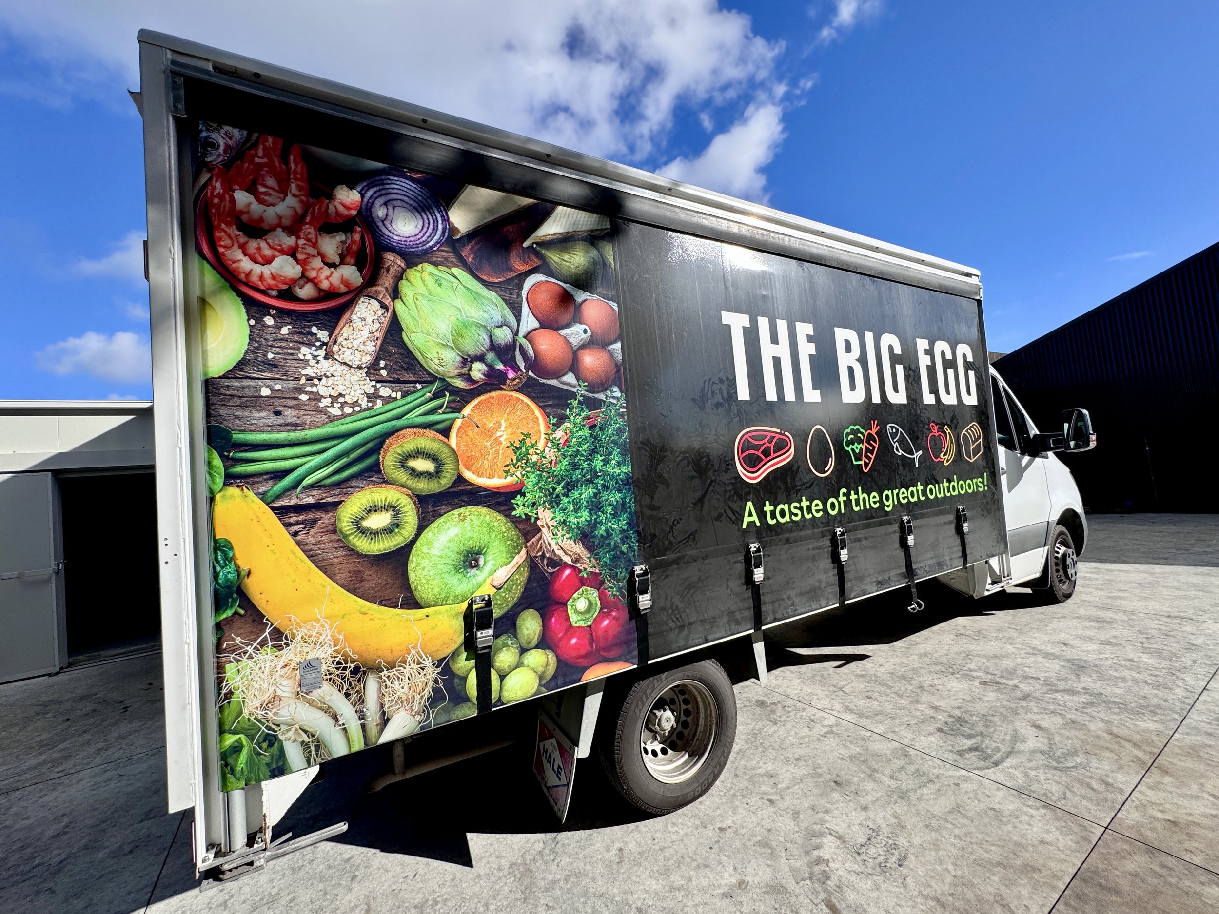 A large, sign-written food truck. The signage on the sides of the truck features a food photo montage wrapped around the back, with the Big Egg logo and its colourful food icons on a black background towards the front.