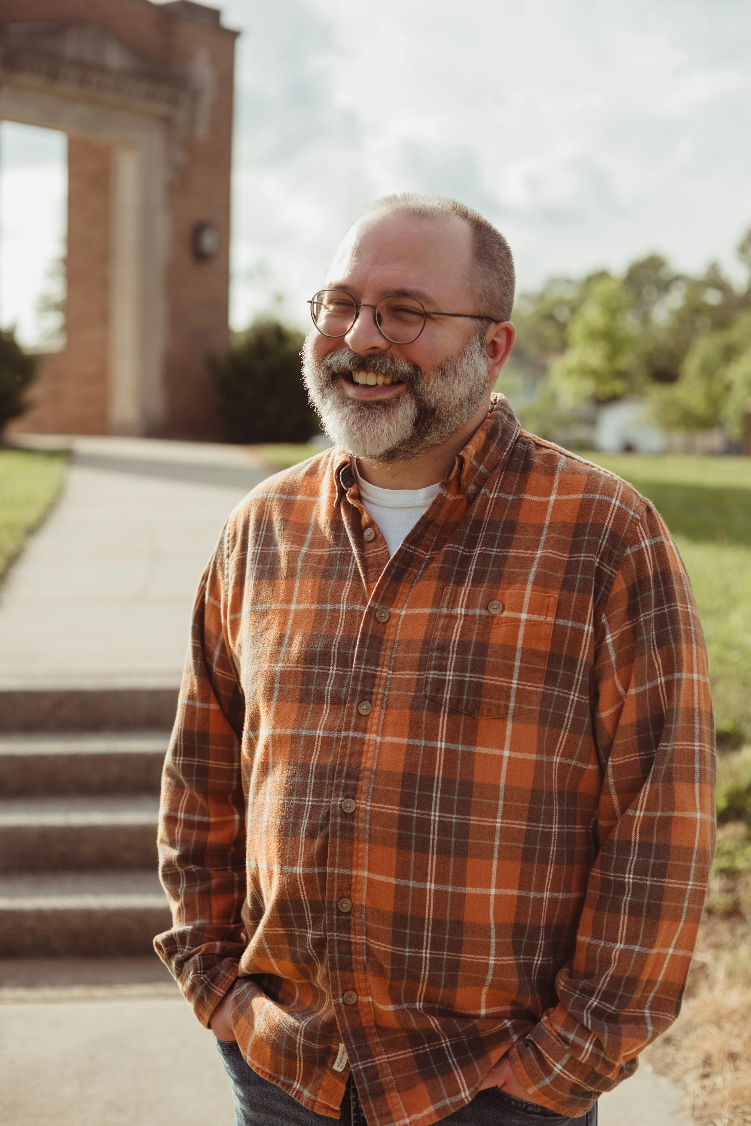 Derek Camp dressed in a plaid shirt stands outdoors with a brick building and stairs in the background.
