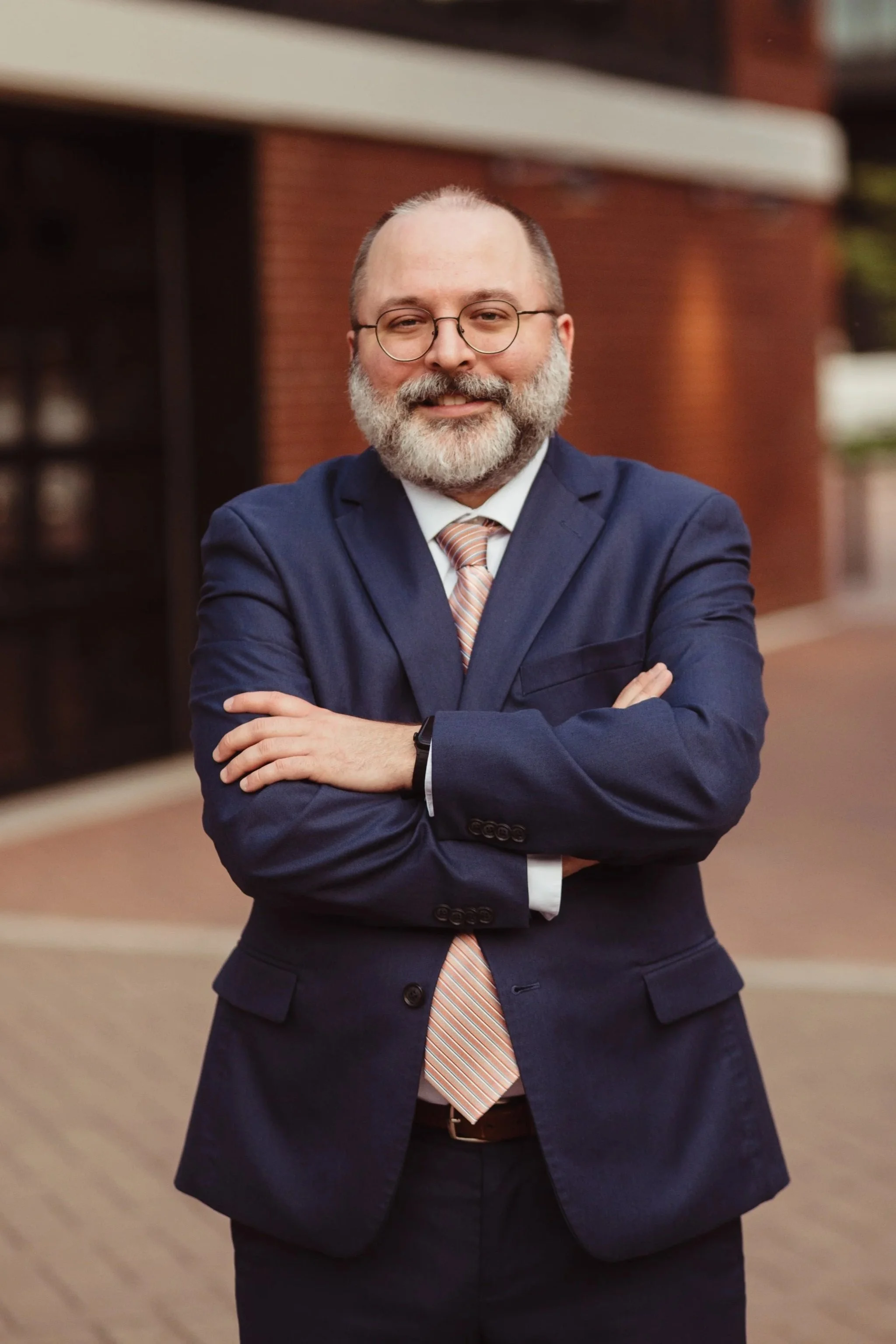 Derek Camp in a blue suit with arms crossed, standing outdoors in front of a brick building.