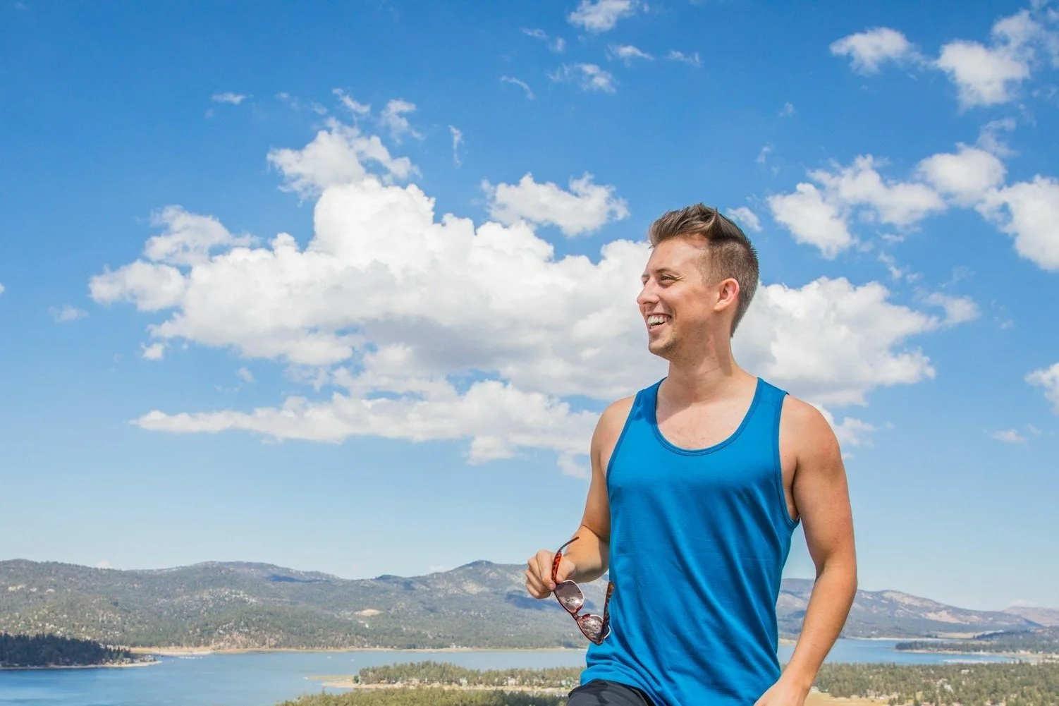 Smiling man in blue tank top standing outdoors with lake and mountains in the background