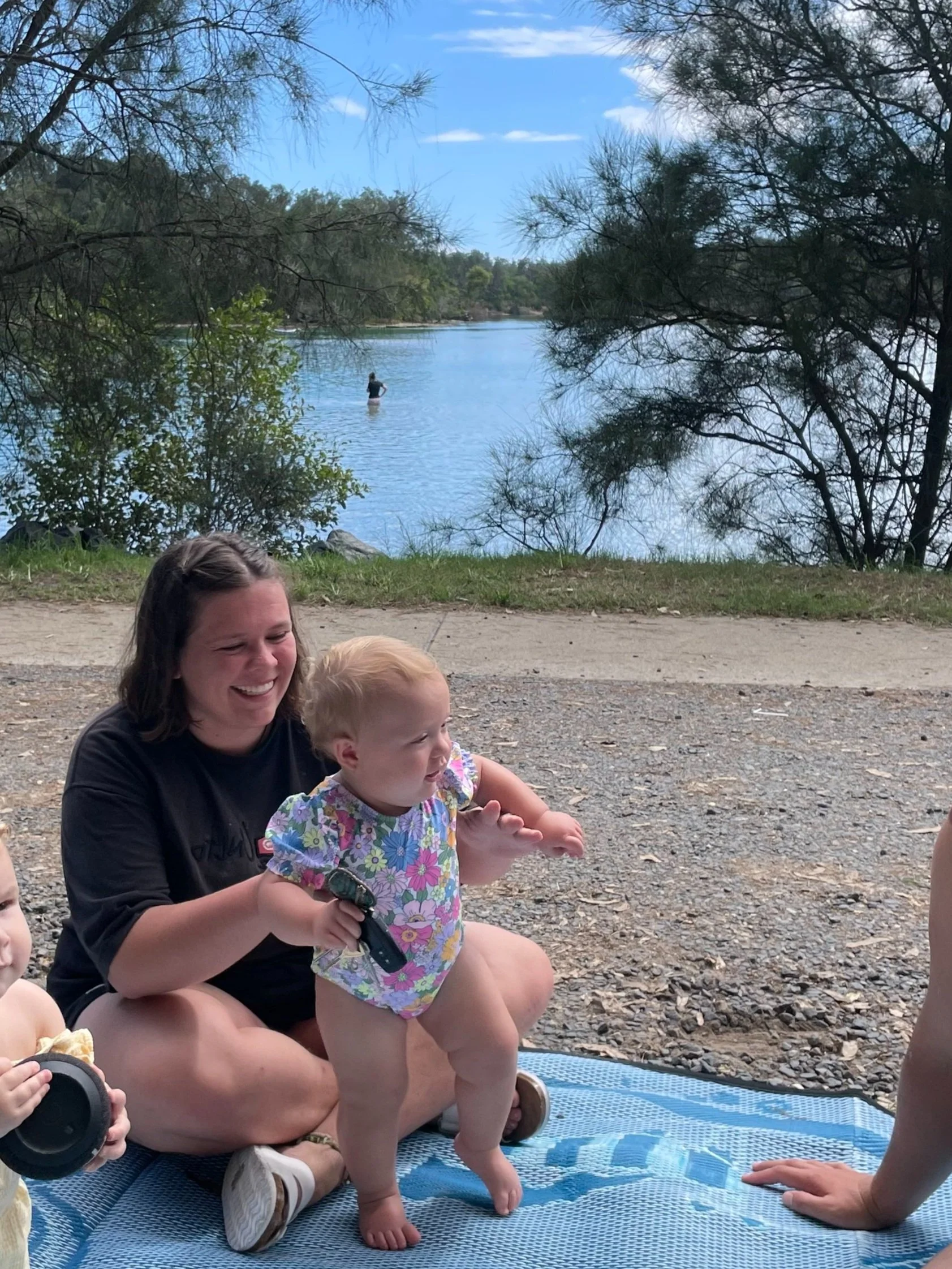 Woman sitting on a mat by a lake holding a standing baby in a floral outfit with trees in the background.