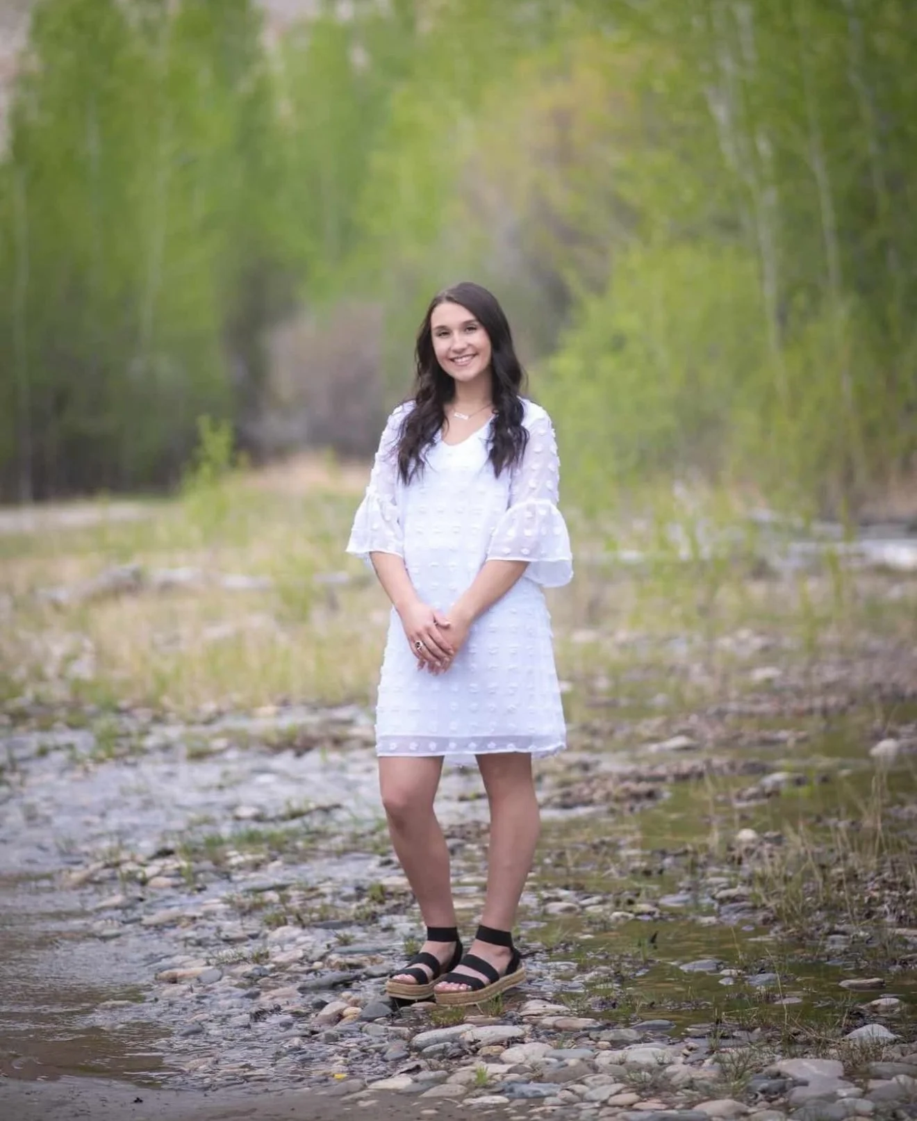 Young woman standing on a rocky creek bed in a forested area with green trees in the background, wearing a white dress and black sandals, smiling at the camera.