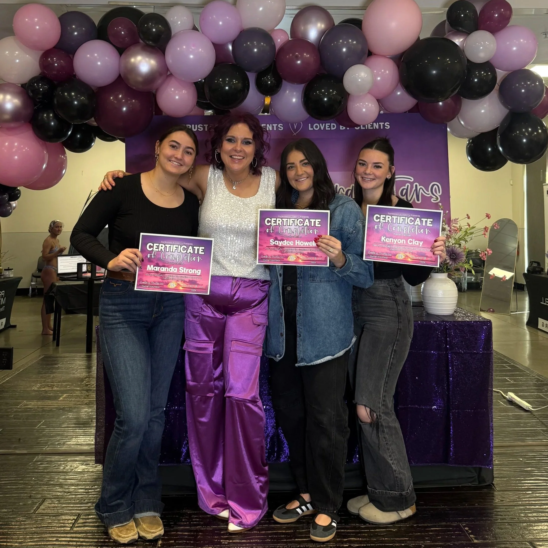 Four women standing together holding certificates with a purple and pink balloon backdrop, celebrating a graduation or certification event.
