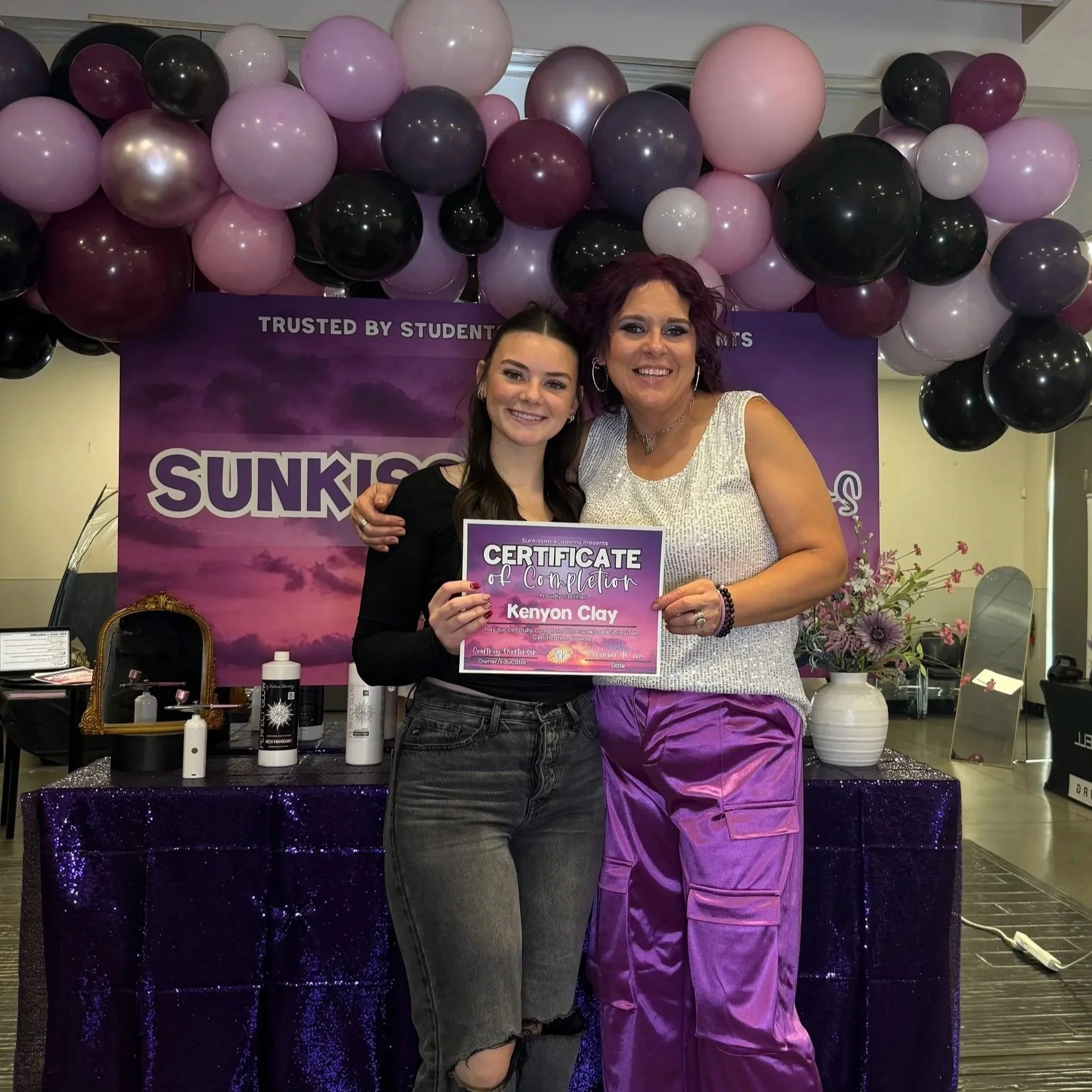 Two women smiling and holding a certificate at a graduation event, with purple and pink balloons and a purple banner in the background.