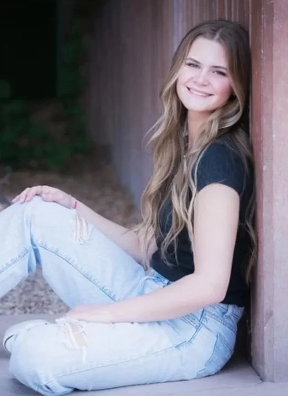 A young woman with long wavy hair sitting and leaning against a wooden wall, smiling at the camera, wearing a black t-shirt and ripped light-wash jeans.