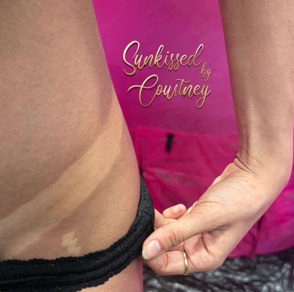 A woman with curly purple hair in a sheer purple lace top and checkered pants uses a photography setup to take pictures of a woman in a bathing suit inside a black tent with instructions displayed on a screen.