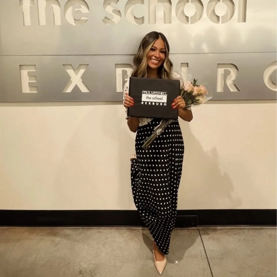 A woman with long wavy hair smiling and holding a bouquet of flowers and a black diploma cover that says "the school RIXBURG" in front of a metallic sign with cutout letters on a beige wall.