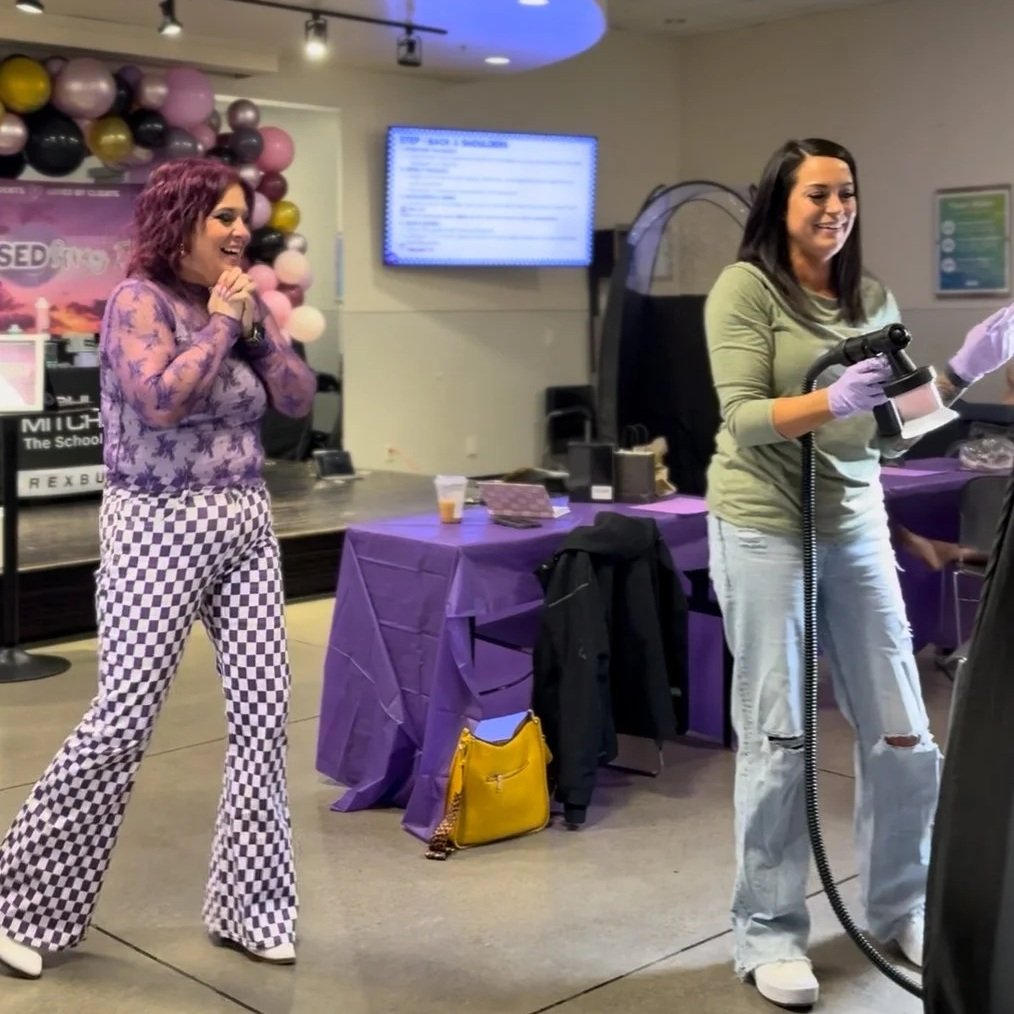 Two women smiling and enjoying themselves at an indoor event, one clapping and the other operating a steam cleaner or carpet cleaner. The background includes balloons, a table with a purple tablecloth, and a large screen.