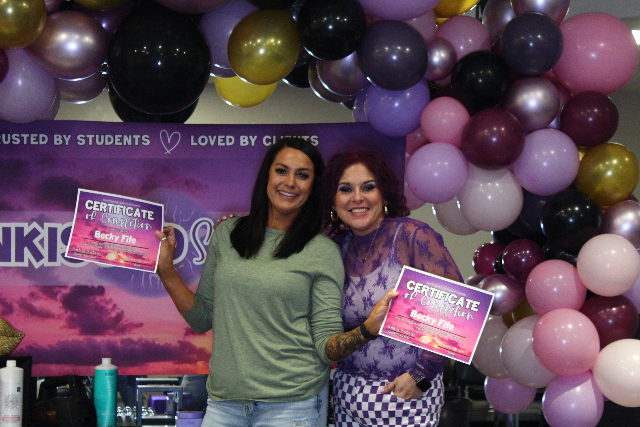 Two women holding certificates of completion at a celebration event, with purple, pink, black, and gold balloons and a purple backdrop behind them.