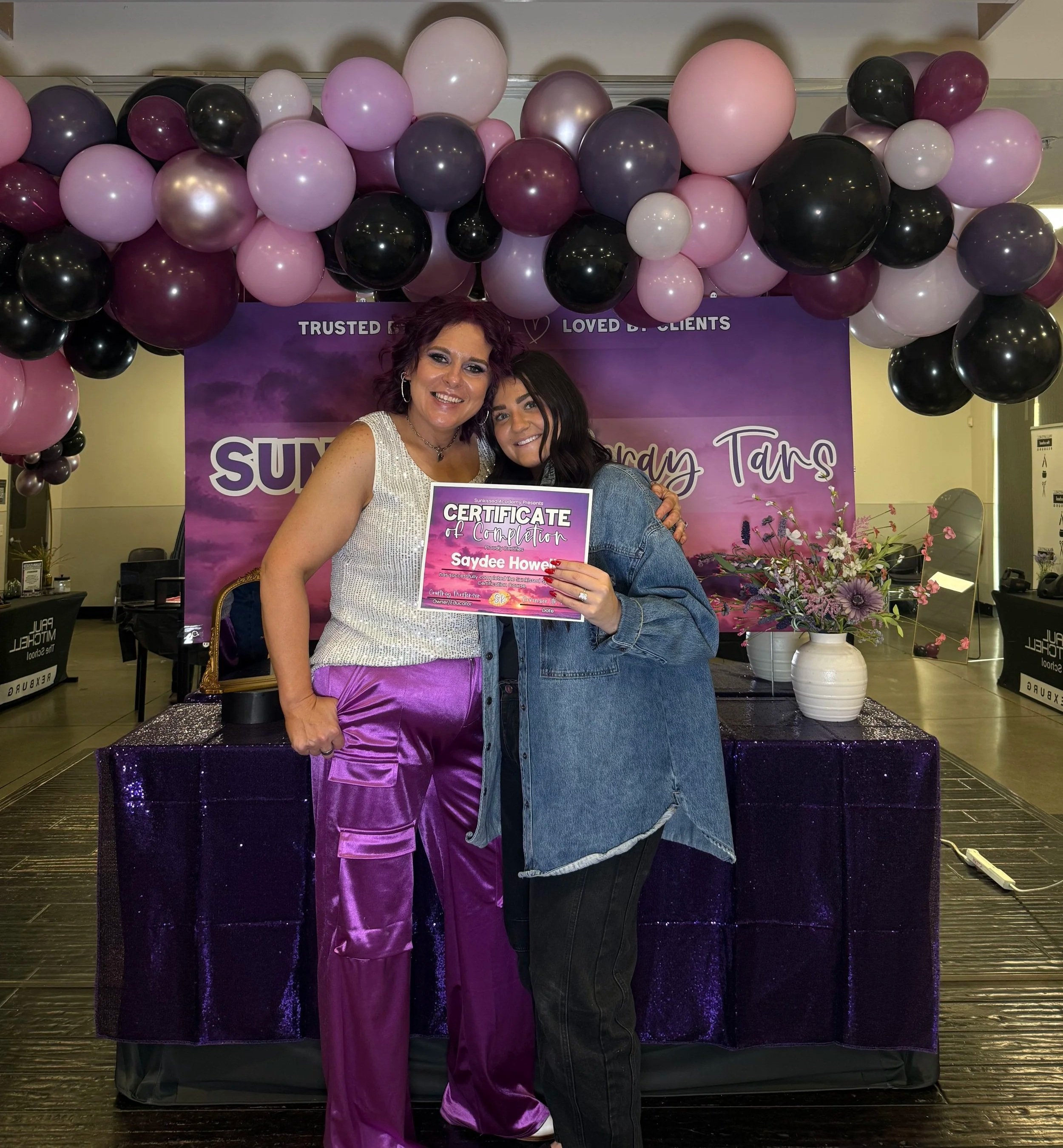 Two women smiling and holding a certificate in front of a purple backdrop and a balloon arch in pink, black, purple, and white. One woman is wearing a shiny silver top and purple satin pants; the other in a denim jacket. Flowers are on a table to the right.