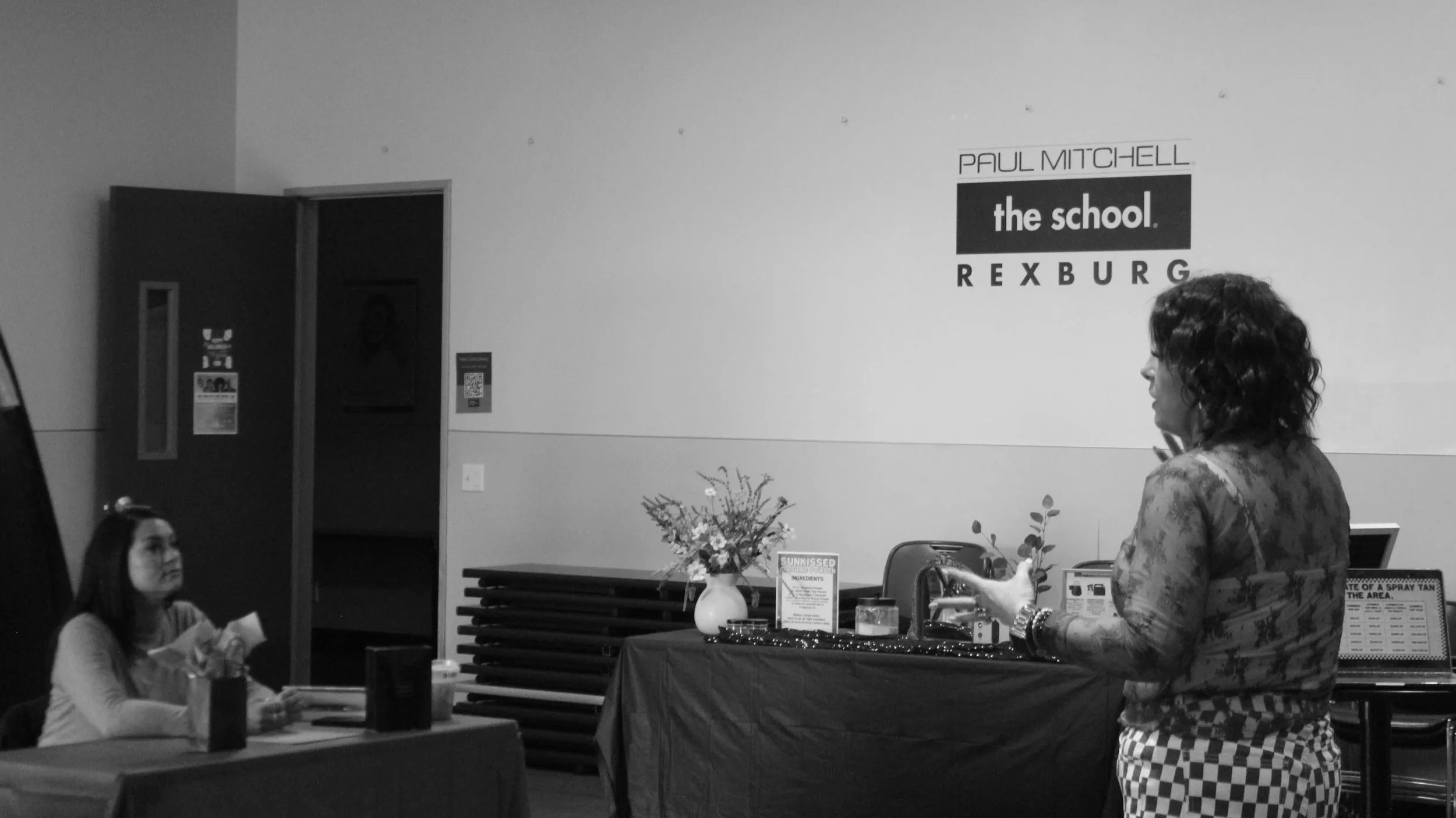 A woman speaking at an event with the sign 'Paul Mitchell the school, Rexburg' on the wall behind her, and a young girl sitting at a table listening.