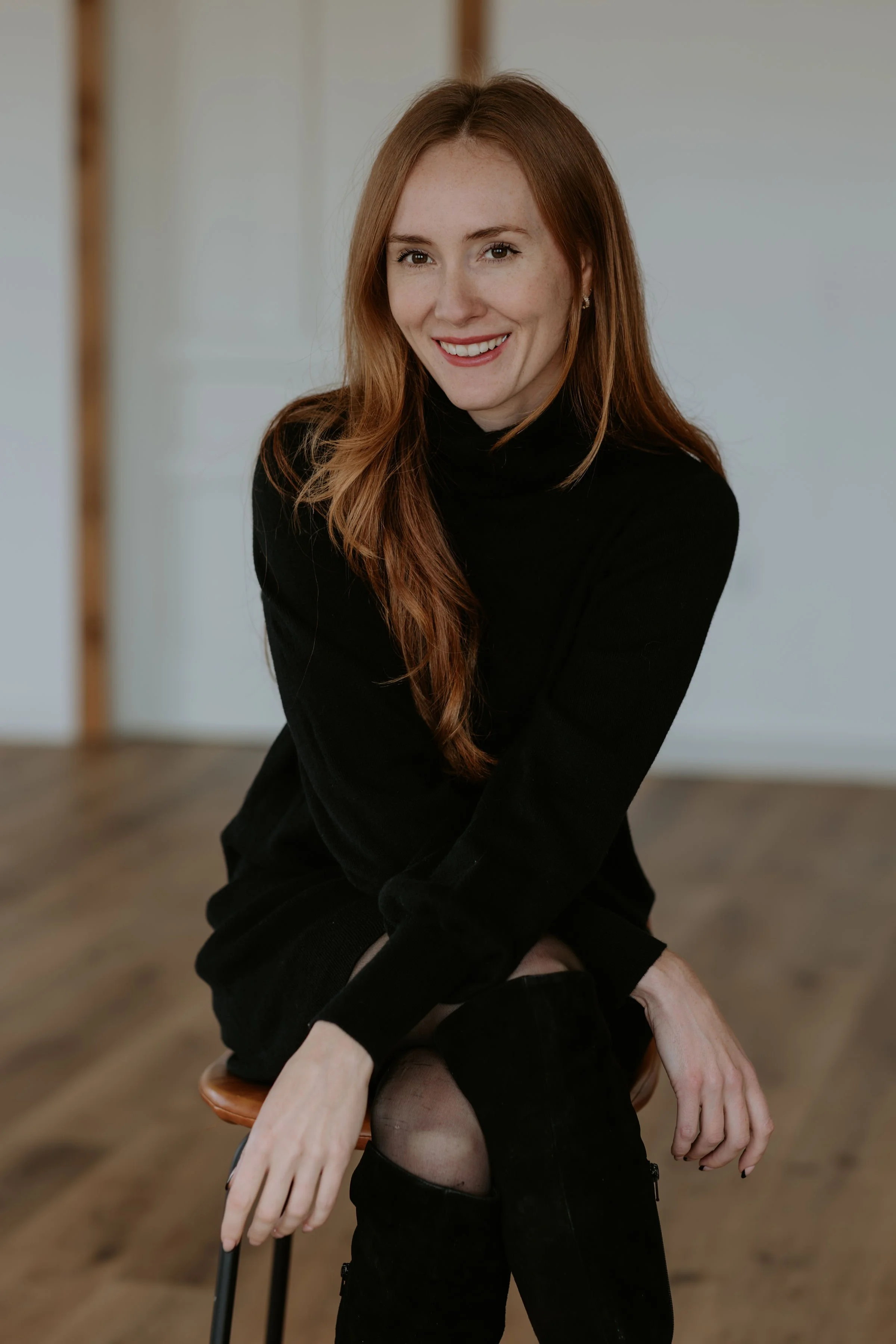 A young woman with long red hair, wearing a black turtleneck, sitting on a stool in a room with wooden floors and a neutral background, smiling at the camera.